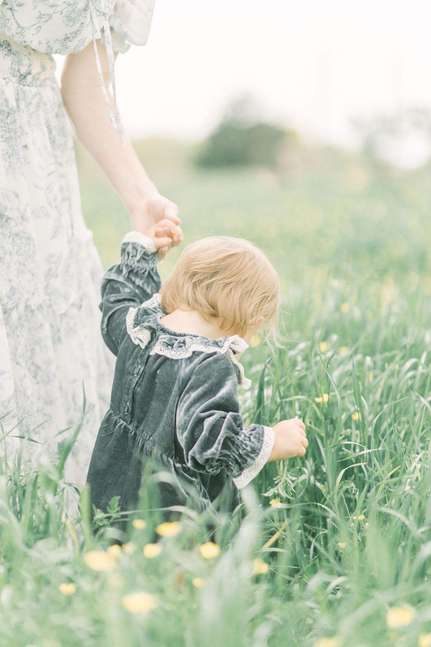 A toddler in a green dress holds mother's hand while walking through a sunny wildflower meadow.