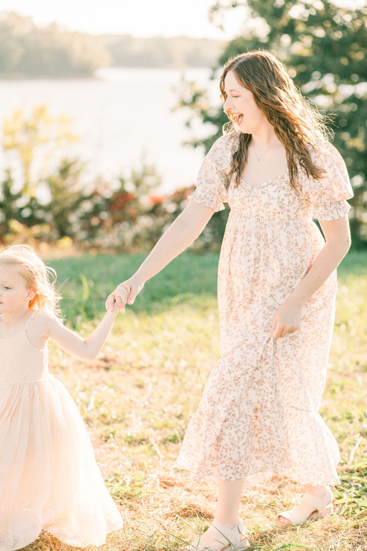 Smiling pregnant woman in floral dress holds hands with toddler girl in pink dress outdoors near a lake.
