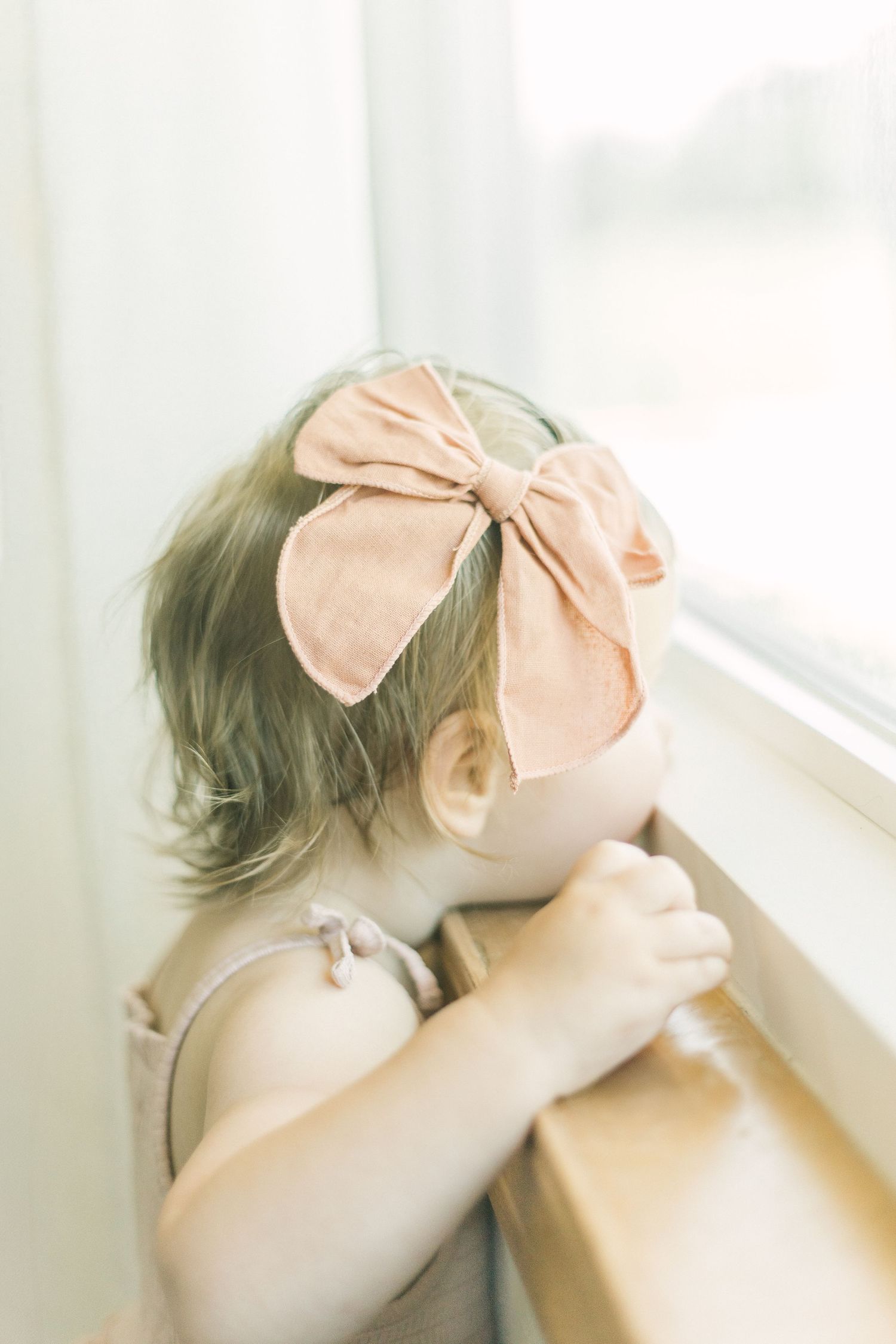 A small child with a peach-colored bow headband peers out a bright window in a soft dreamy scene in Springfield, Missouri.