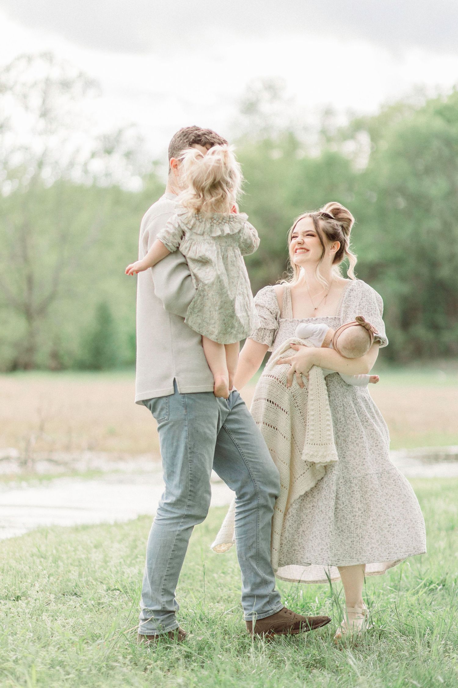 A joyful family poses outdoors, father holding toddler girl, mother cradling newborn, wearing soft neutral tones in a green field.