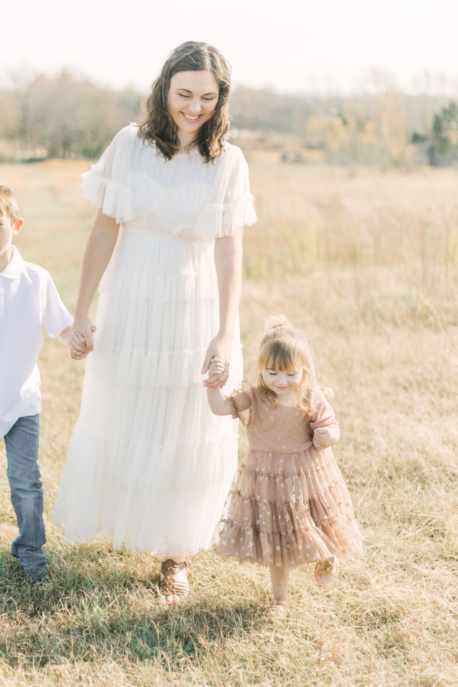 A smiling mother in a white dress holds hands with her two young children while walking through a sunny golden field.