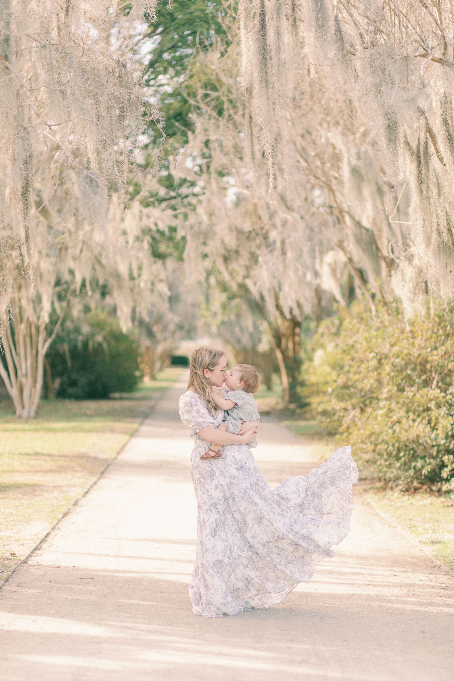 A dreamy motherhood session by a Springfield, MO photographer. A mother twirls in a flowing dress, holding her baby under Spanish moss trees
