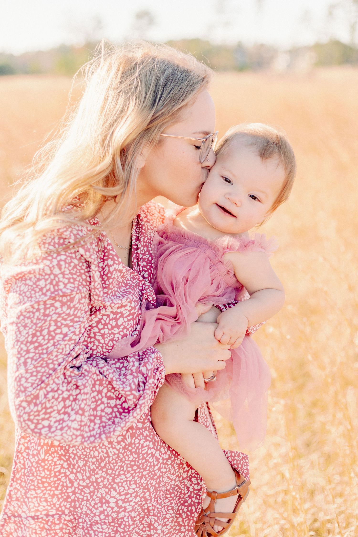 Mom kisses baby girl in pink dress during golden hour field session in Springfield, MO. Light and airy family photo.