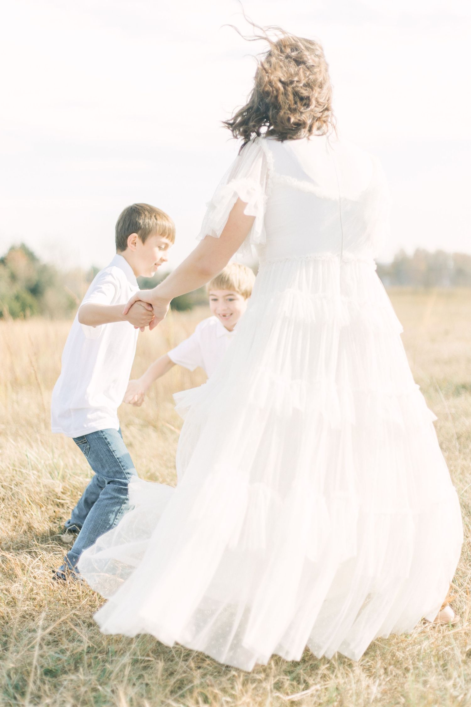 Springfield, MO family photographer captures a joyful moment of a mother playing Ring Around the Rosie with her sons in a sunlit field.