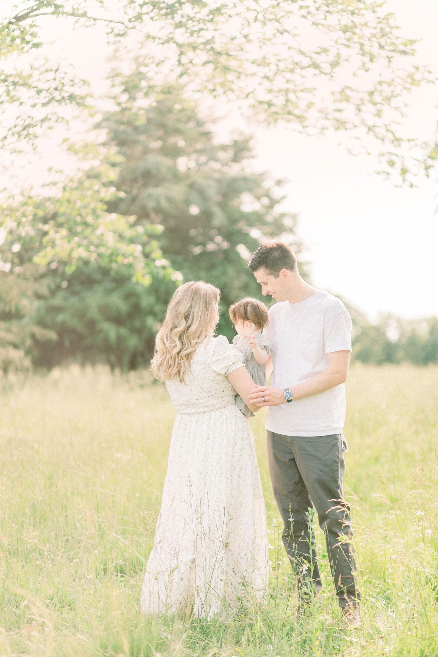 Family stands together in a sunlit meadow wearing white and casual clothing during a dreamy outdoor photo session in Marshfield, Missouri.