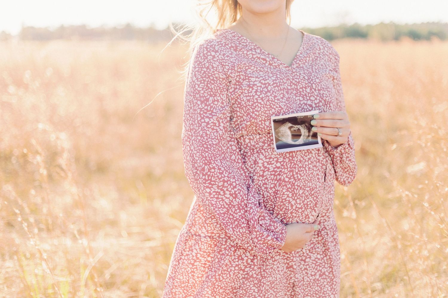 A pregnant woman in a soft pink floral dress stands in a field at golden hour, gently cradling her baby bump with one hand, by Marshfield Mo