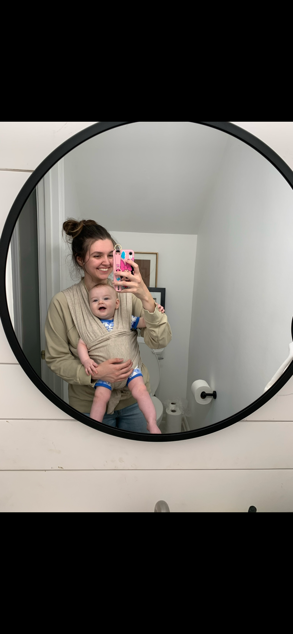 A mirror selfie showing someone in a beige sweater holding a baby in a bathroom with a round black-framed mirror in Springfield, Missouri.