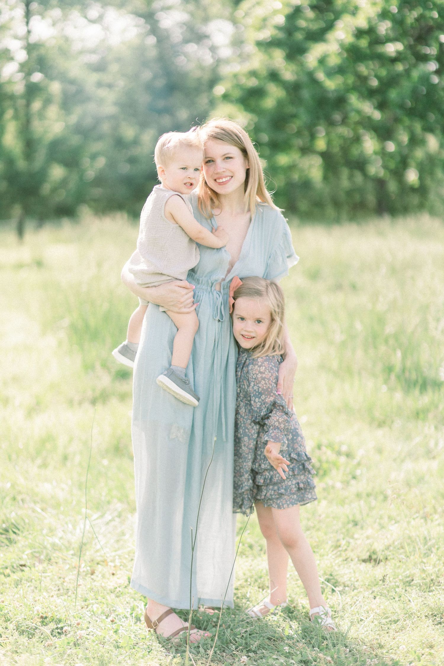 Mother in flowing blue dress embraces two children while standing in a sunlit field at Fellows Lake in Springfield, Missouri.