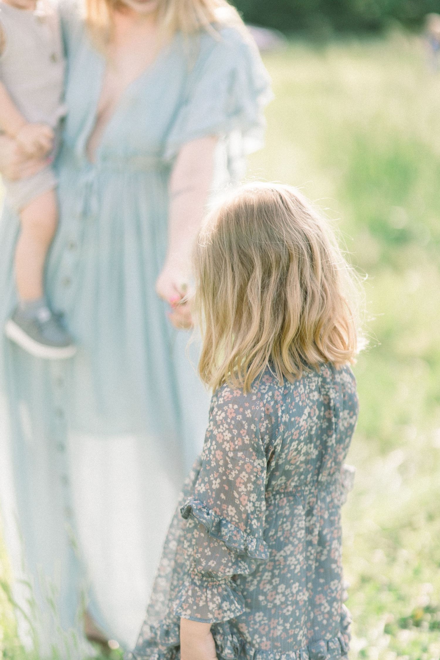 A young girl in a floral dress stands in a sunny Springfield, Missouri meadow next to her mother wearing a light blue flowing dress.