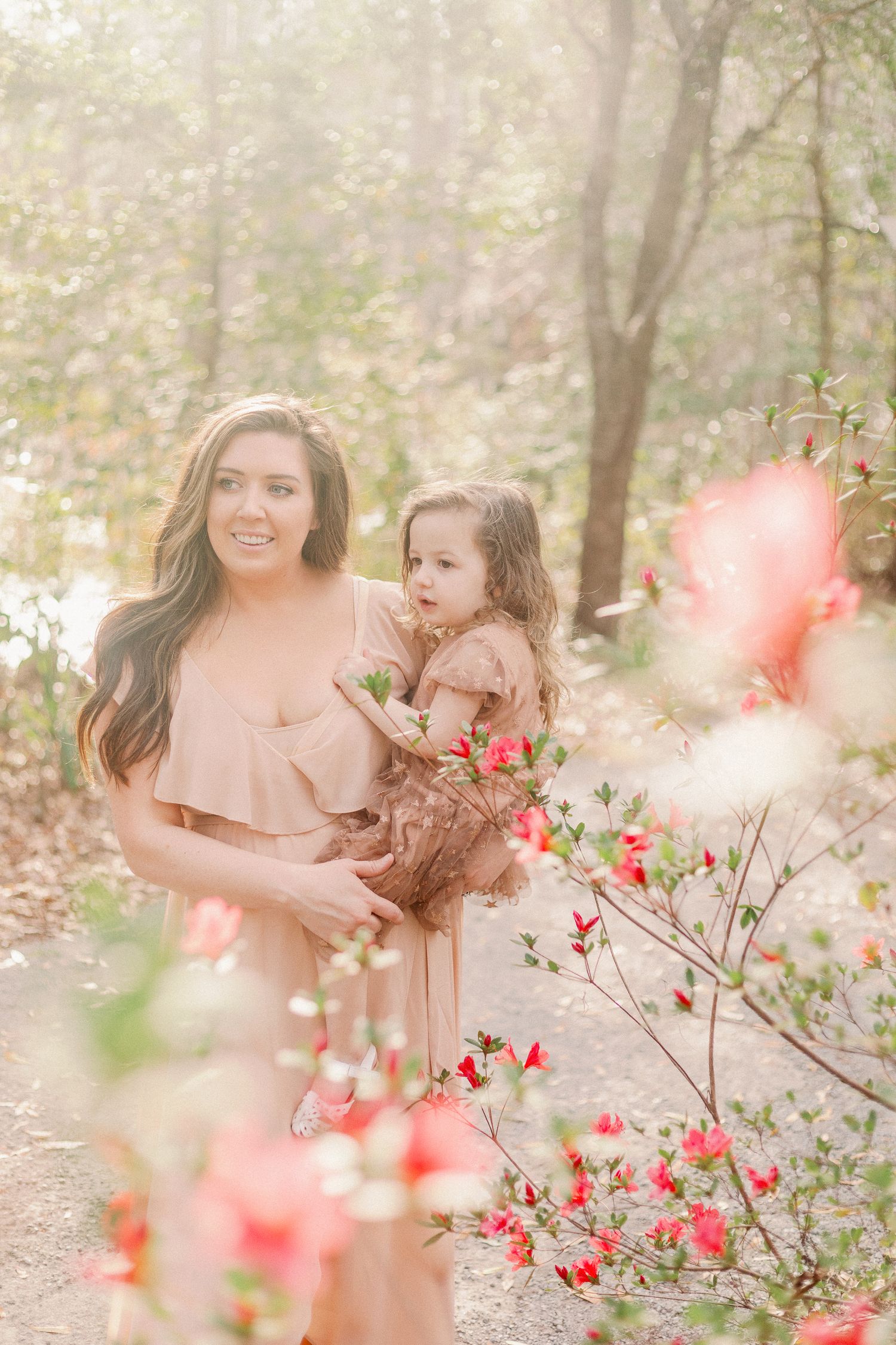 A mother in a blush dress holds her toddler daughter among blooming pink azaleas in a sun-dappled forest.