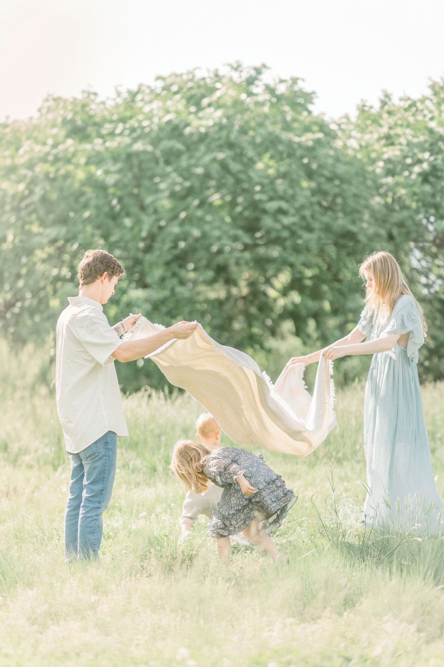 Family plays together in a sunny field, shaking out a beige blanket while a small child ducks underneath in Springfield, Missouri.
