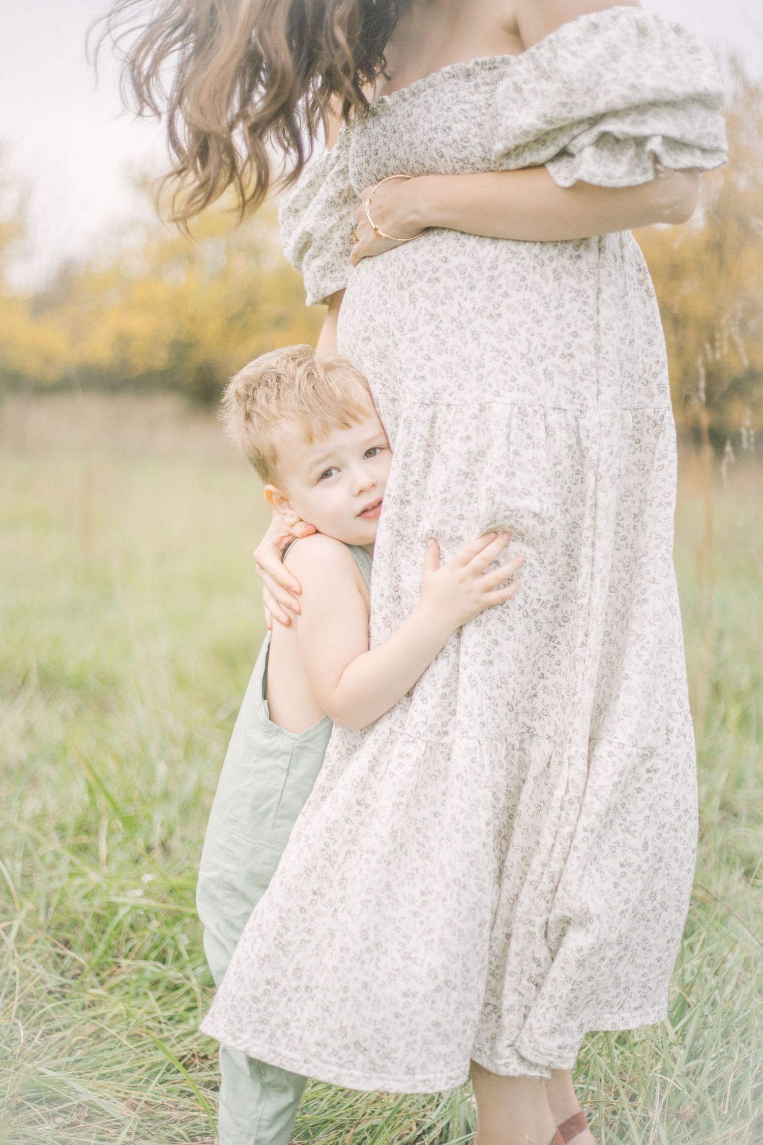 A small boy in green overalls hugs his mama in a flowing floral dress in a sunlit meadow with golden bokeh background in Springfield, MO.