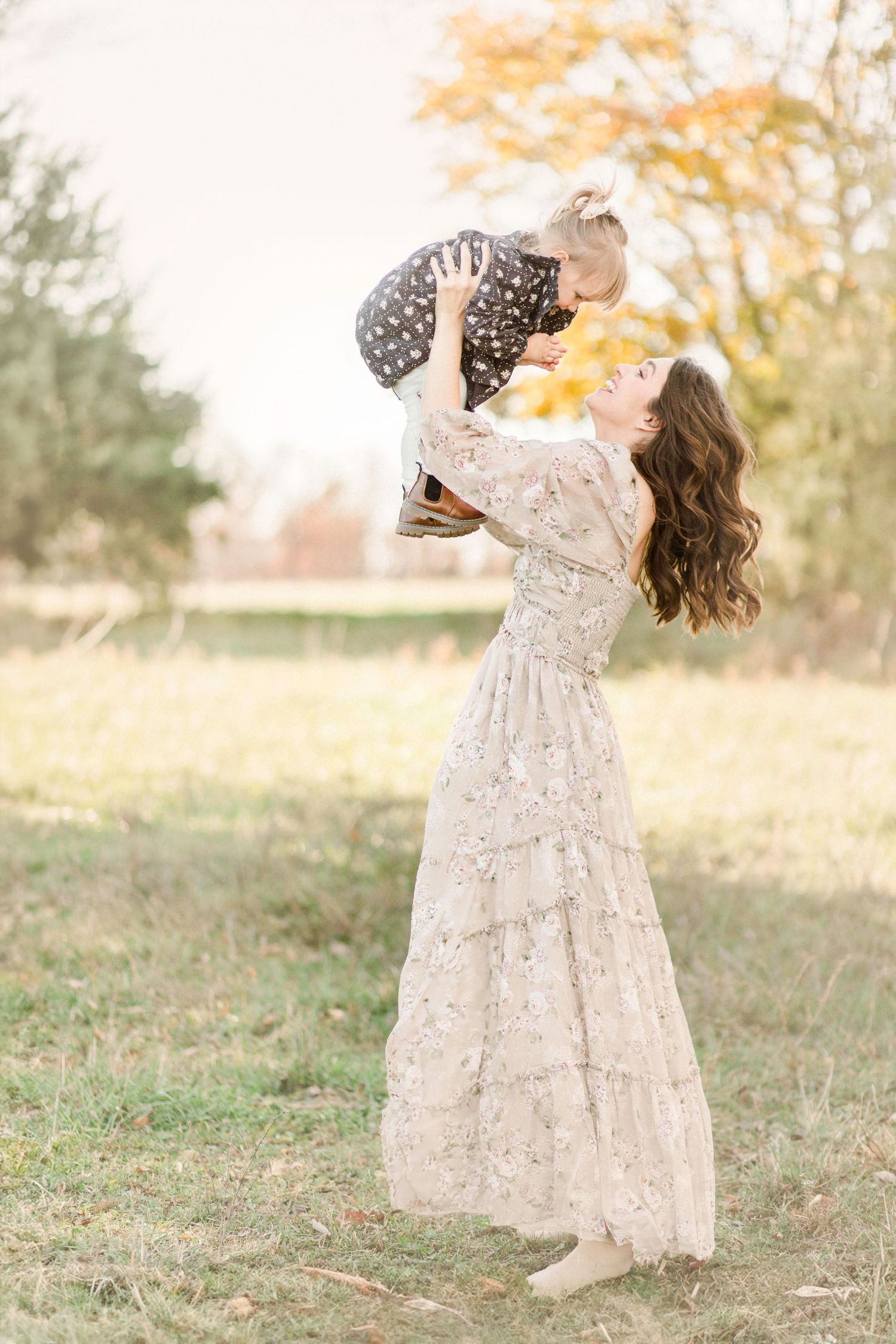 A mother in a floral gown lifts her toddler joyfully in an autumn field.