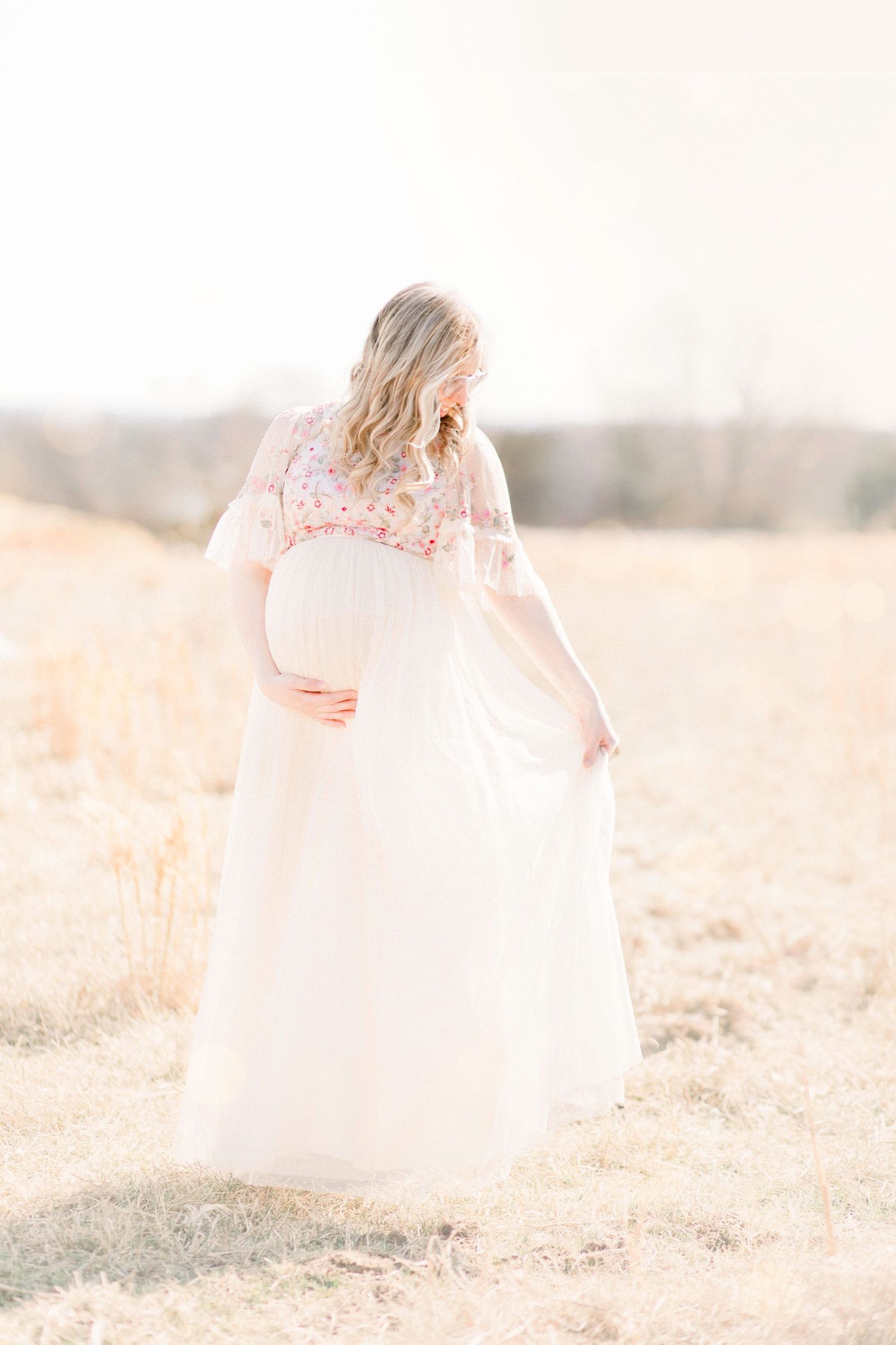 Pregnant woman in white floral dress cradling baby bump in golden field during maternity photo session in Springfield, Missouri.