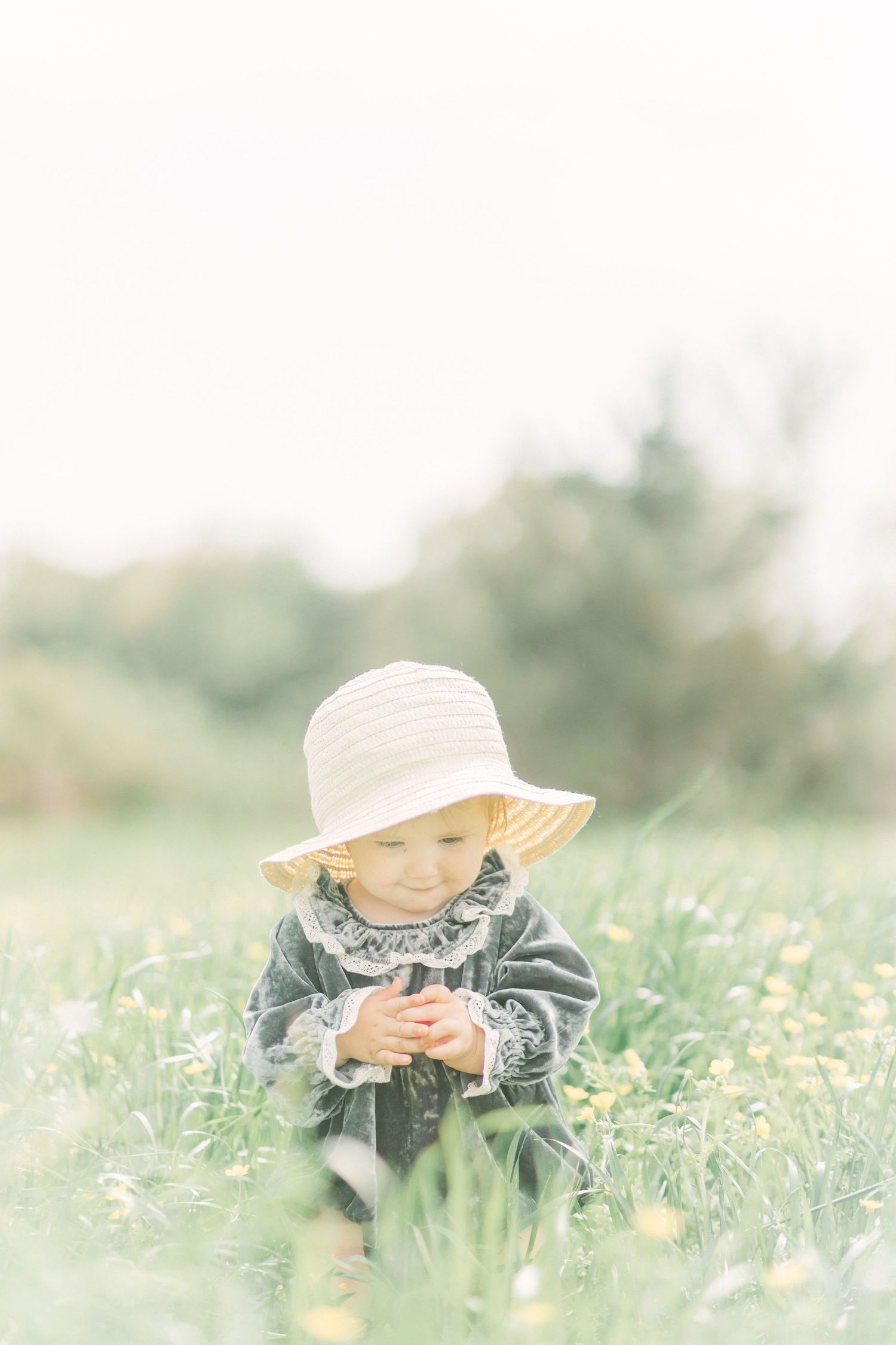 A small little girl wearing a sun hat sits among buttercups in a dreamy Springfield, Missouri meadow on a soft spring day.
