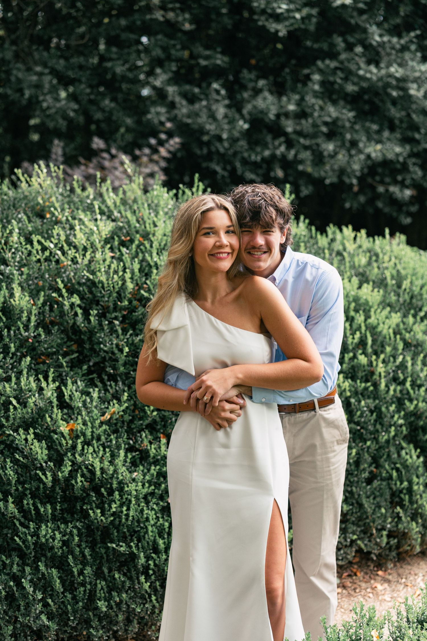A couple in white attire embraces while standing in front of lush green foliage.