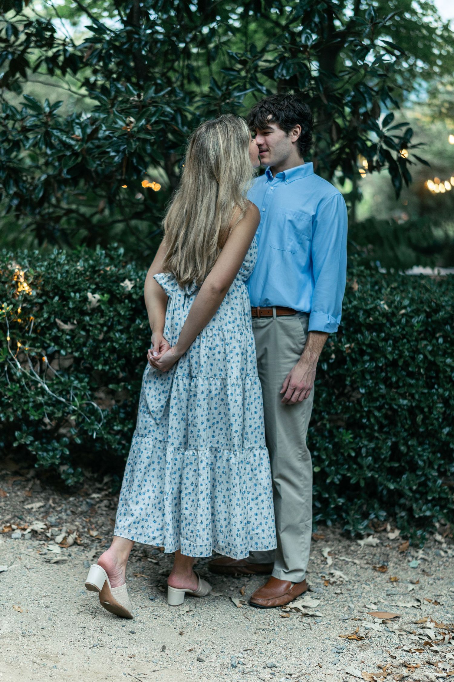 A romantic couple in casual attire pose together against lush greenery and string lights at dusk.