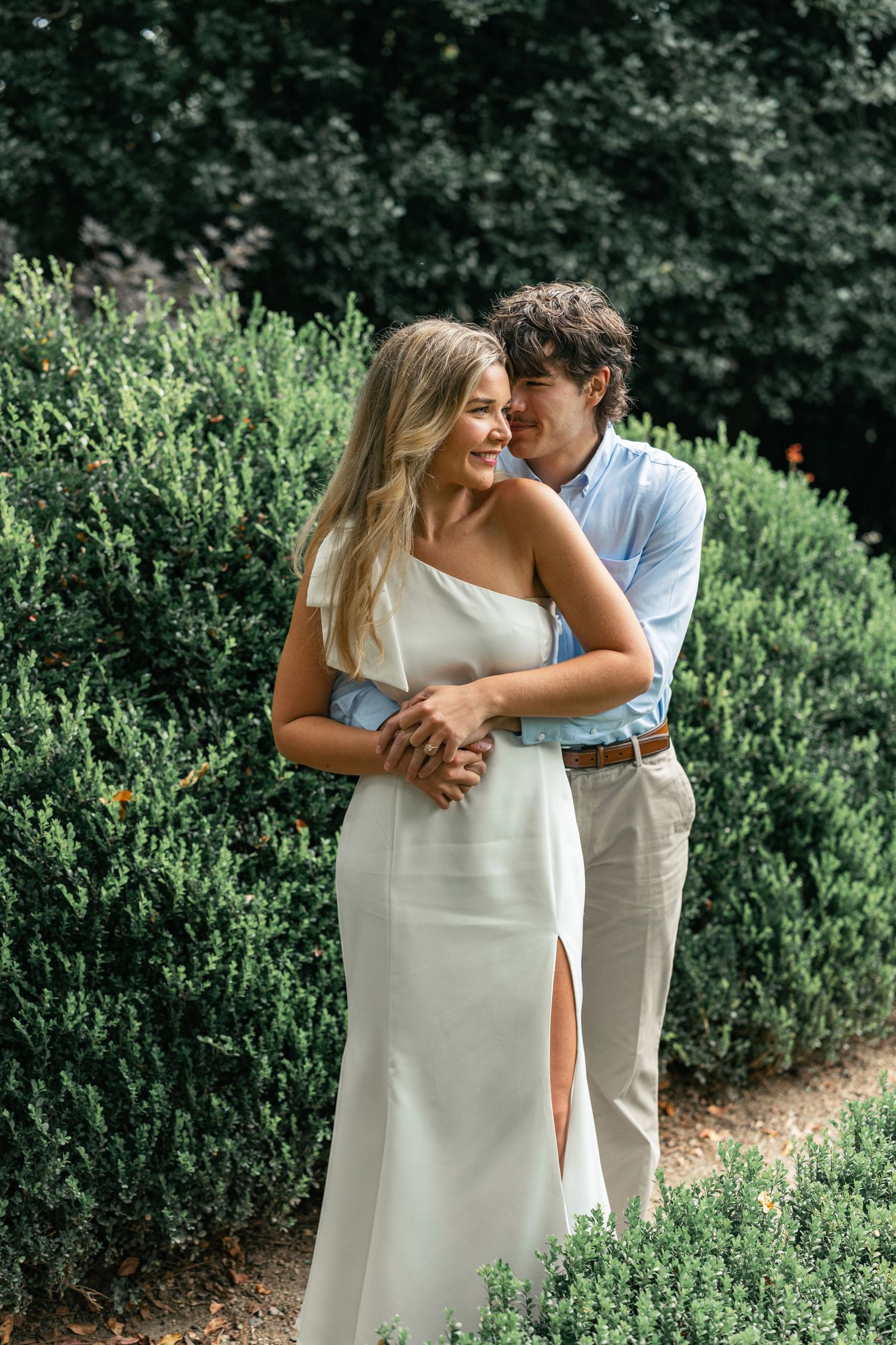 Romantic couple embrace among garden greenery in formal attire.
