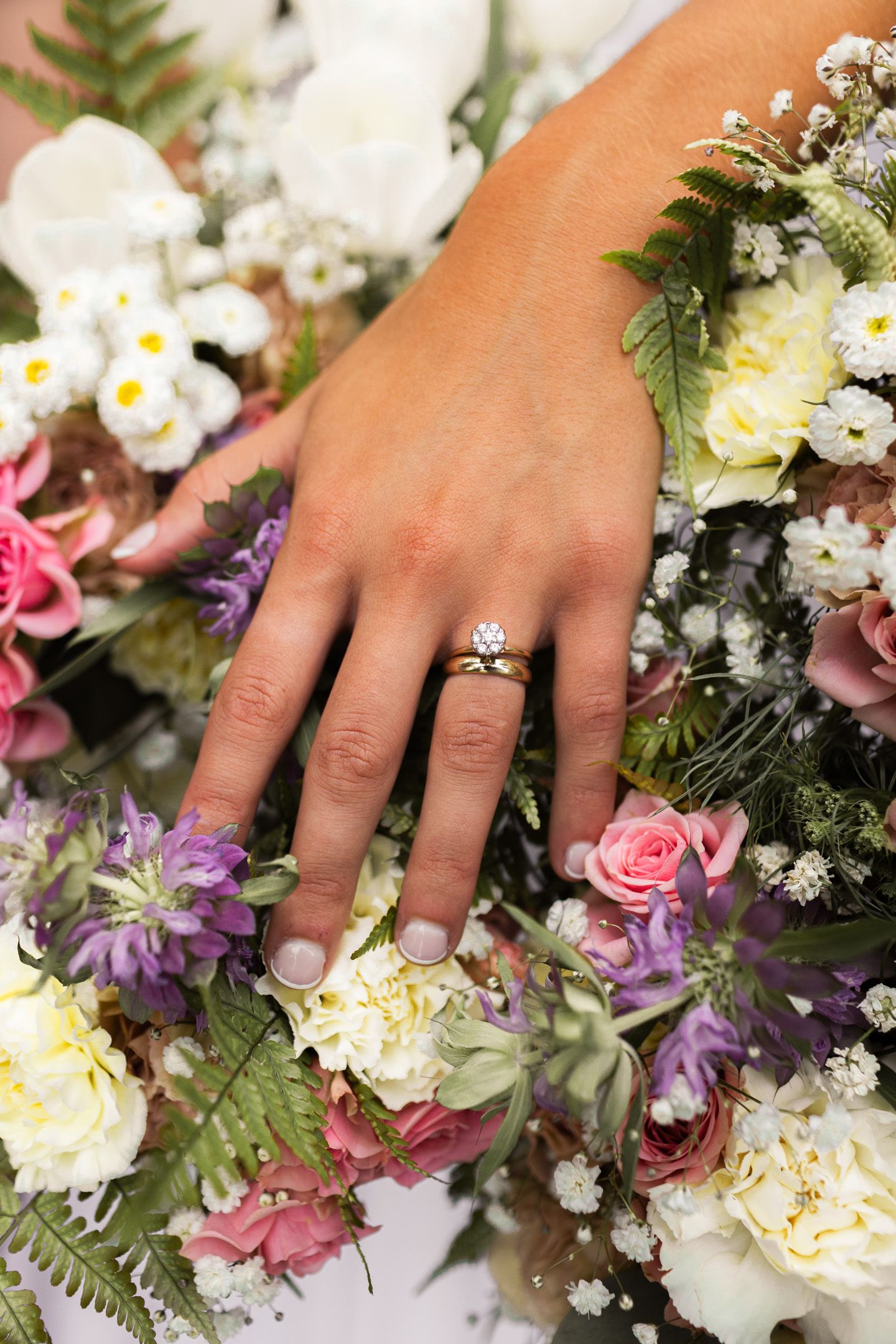 Close-up of engagement ring on hand resting on colorful wedding bouquet with mixed flowers.