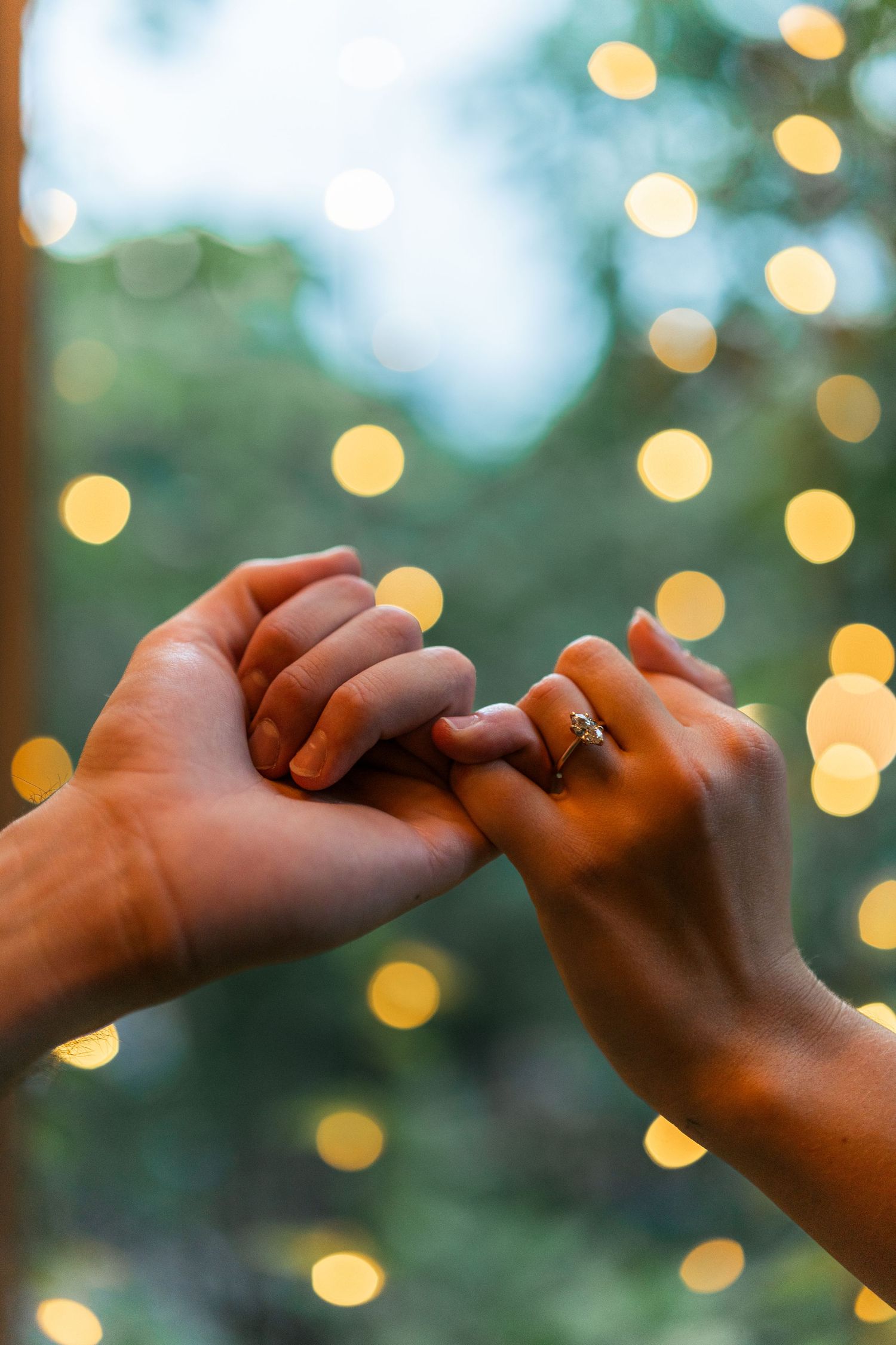 Hands intertwined with engagement ring glowing in evening bokeh lights.