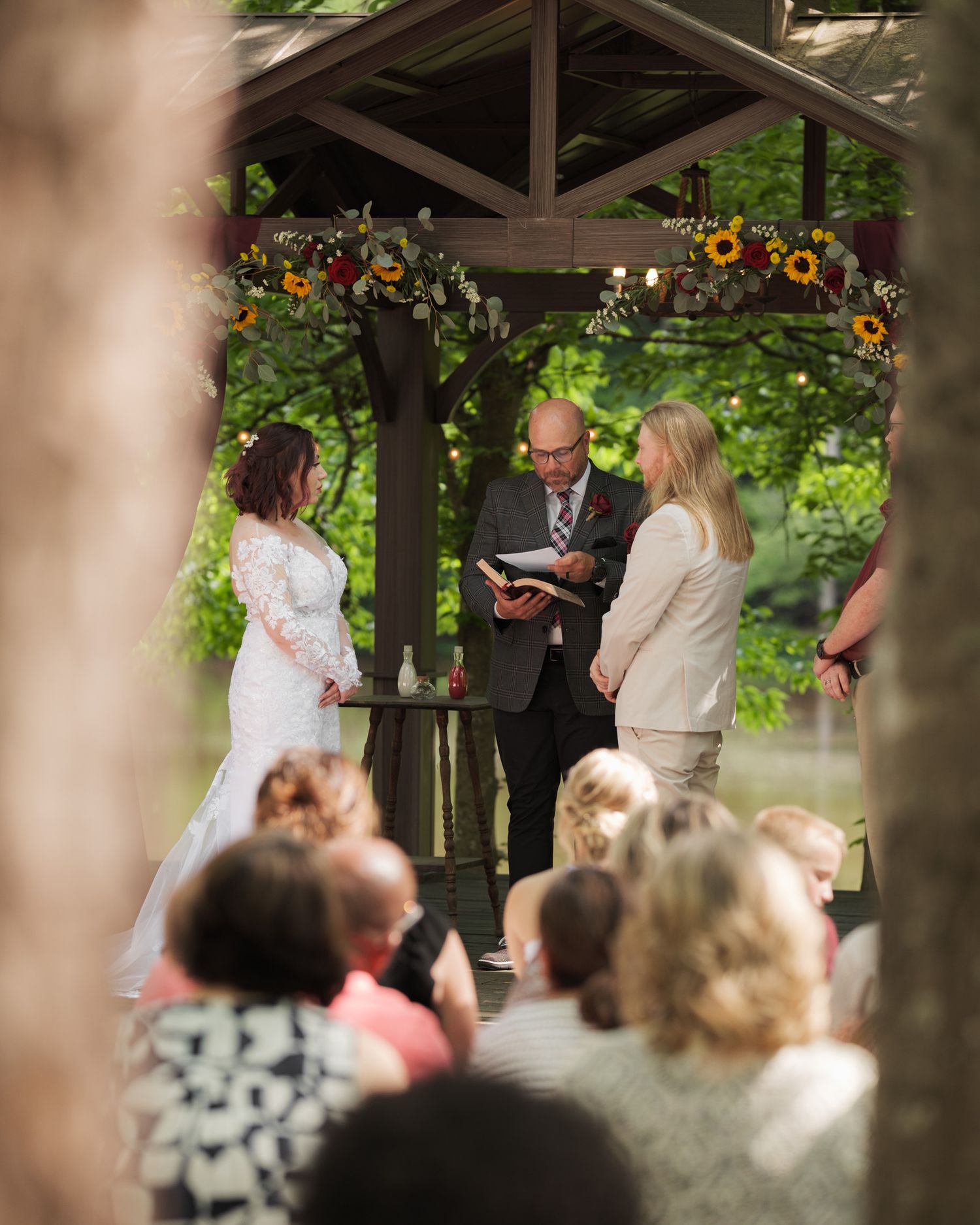 Wedding ceremony taking place under a wooden gazebo surrounded by flowers and guests.