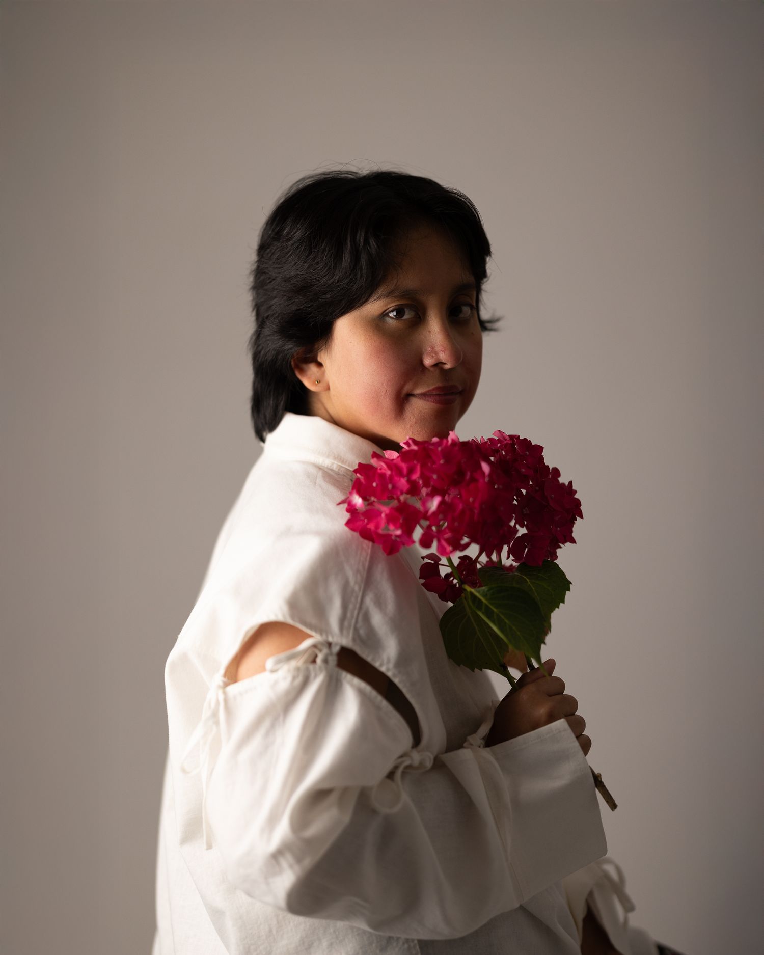 Colour portrait of a woman with short dark hair holding a large pink peony bouquet, looking away, plain background