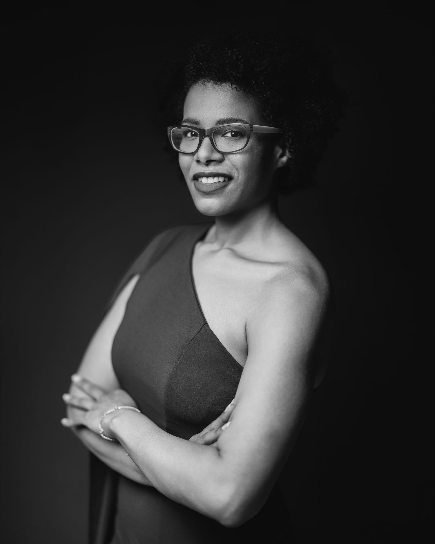 Black and white portrait of a woman with natural hair and glasses in a one-shoulder top, arms crossed, dark studio background