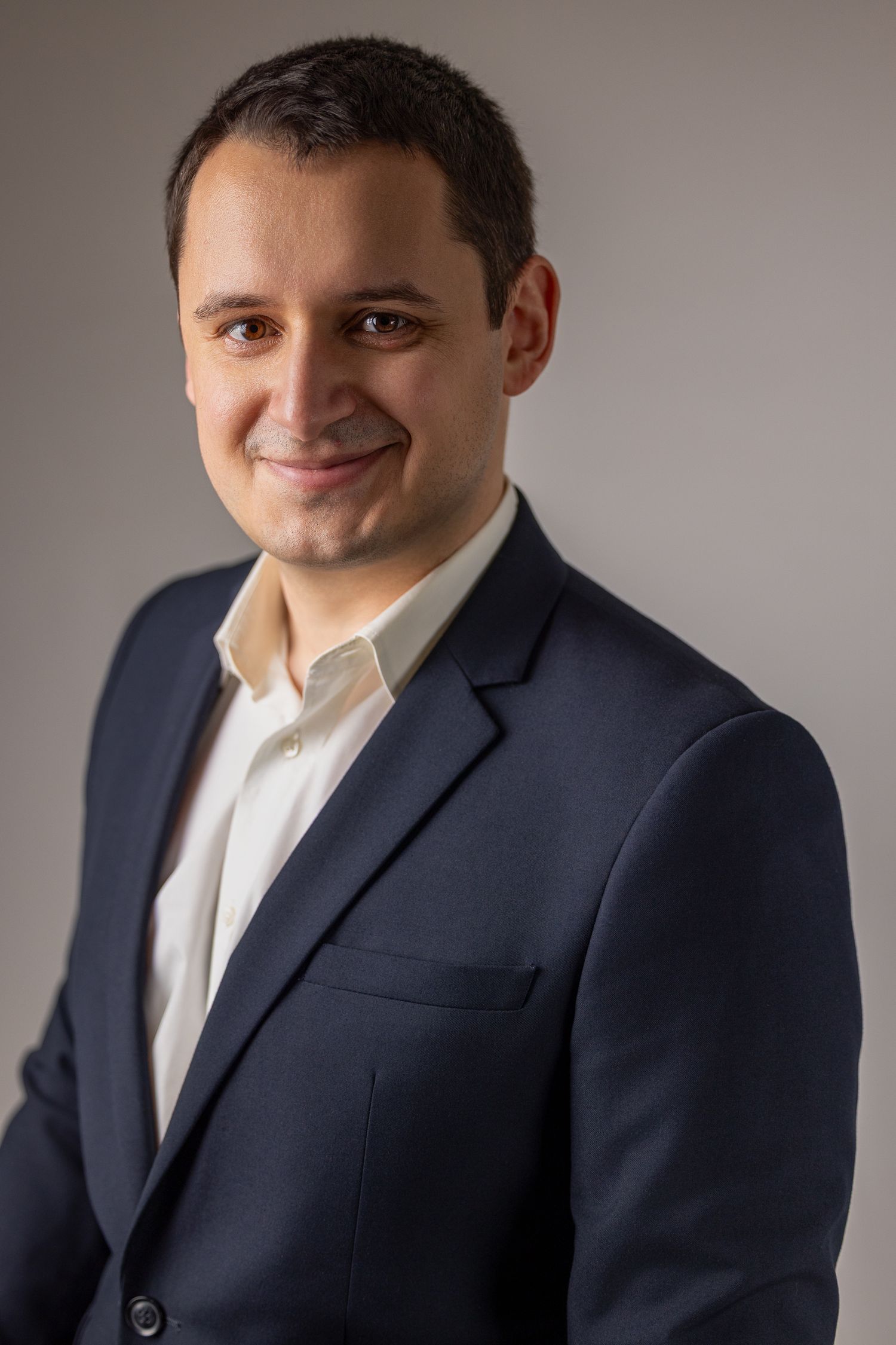 Professional headshot of a man in a navy suit jacket against a neutral grey background