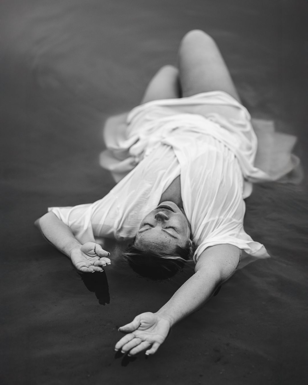 Black and white portrait of a woman in a white dress lying on a dark studio floor, dramatic composed pose