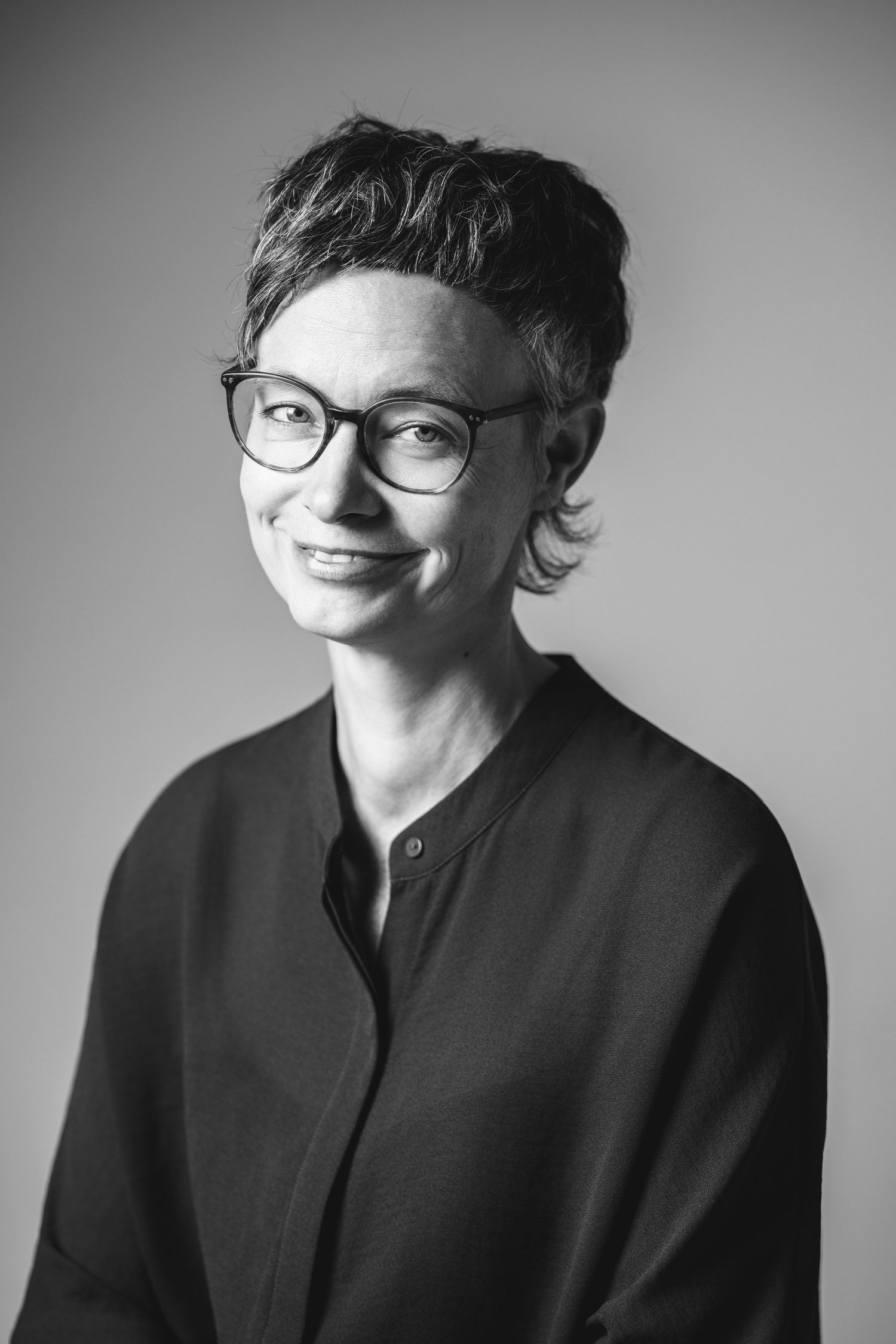 Black and white headshot of a woman with short dark hair and glasses in a dark collarless top, grey background