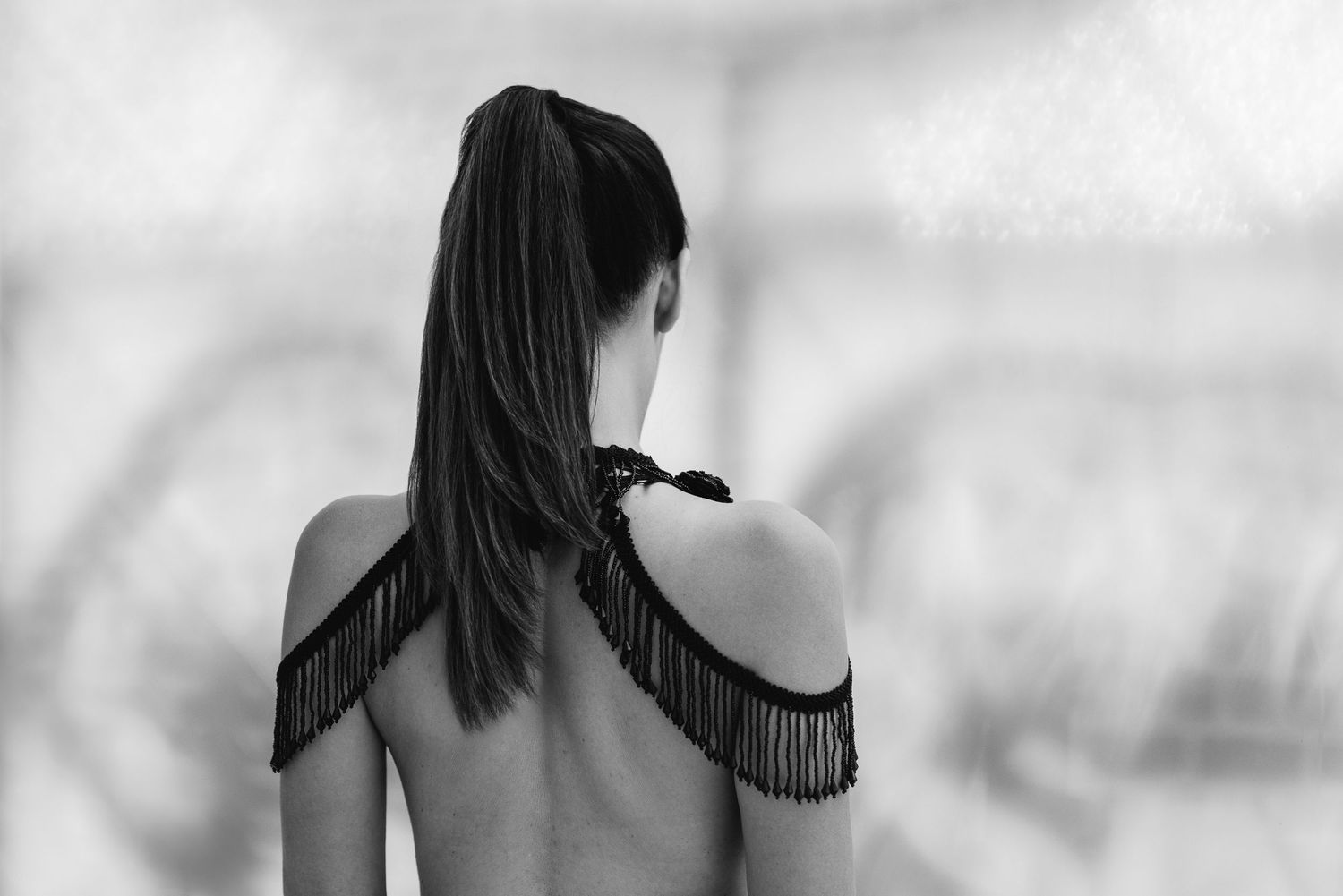 Black and white boudoir portrait from behind, a woman with a long ponytail wearing a fringed shoulder piece, bright soft background