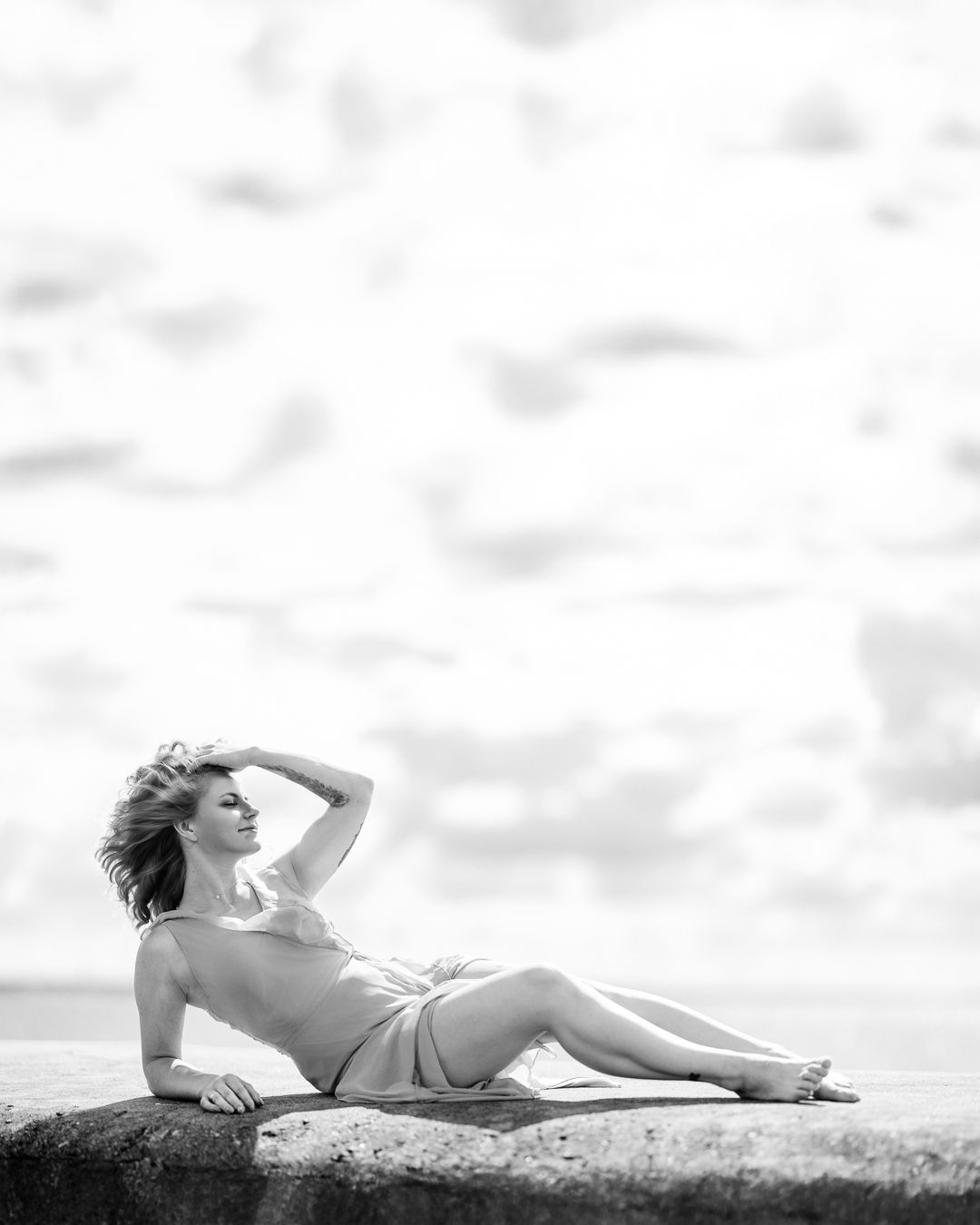 Black and white portrait of a woman lying on coastal rocks, arm raised, against a cloudy sky