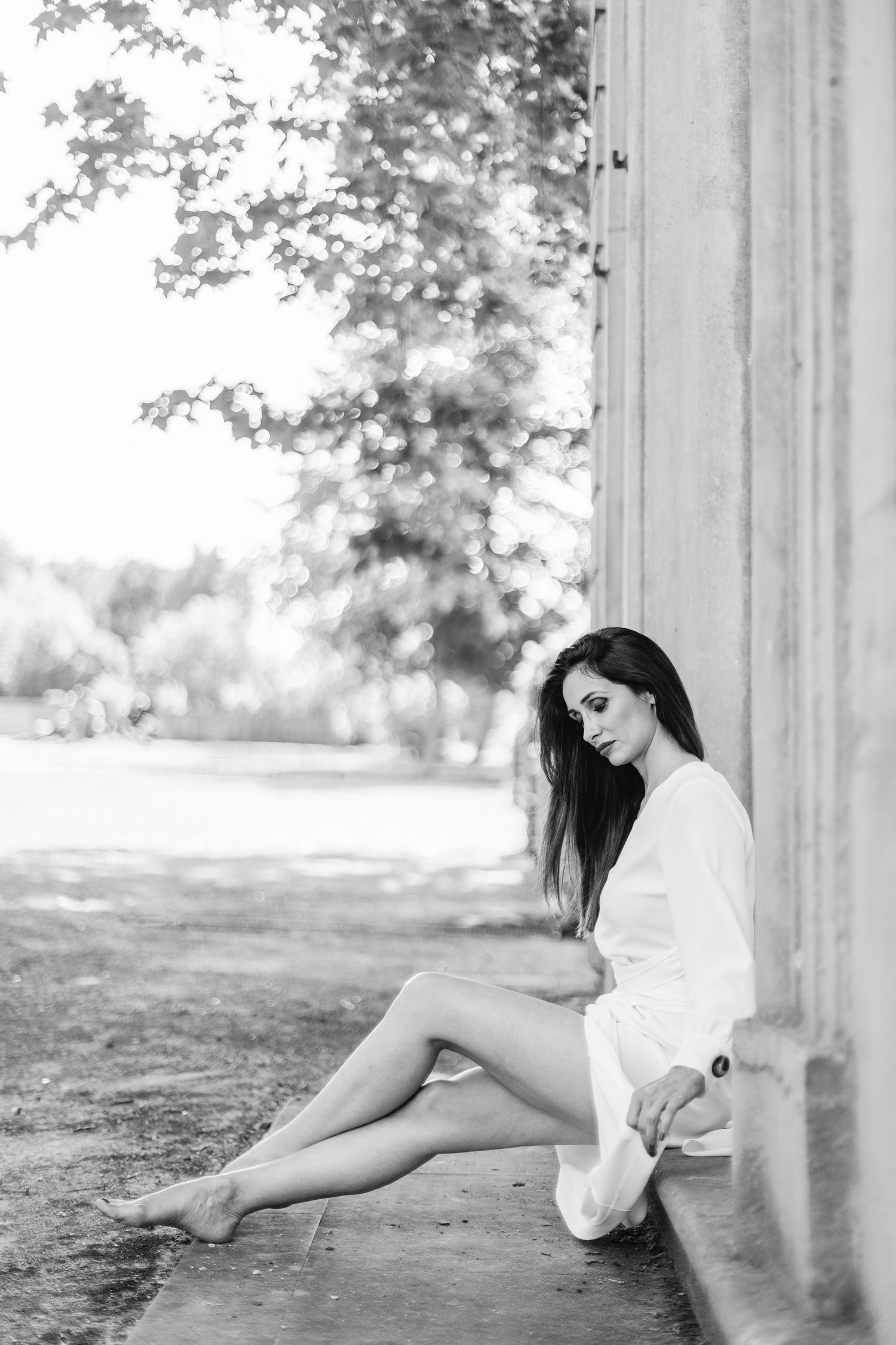 Black and white portrait of a woman in a white shirt seated against a stone column, outdoor setting with trees behind