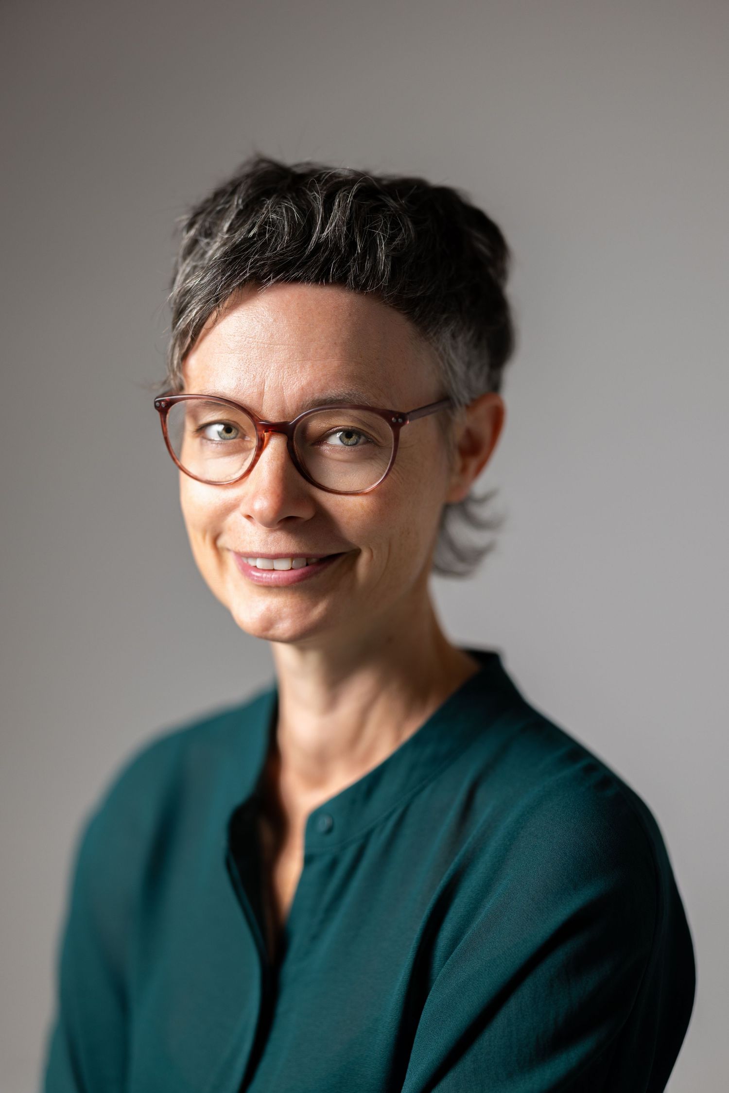 Colour headshot of a woman with short greying hair and glasses in a dark teal top, grey background