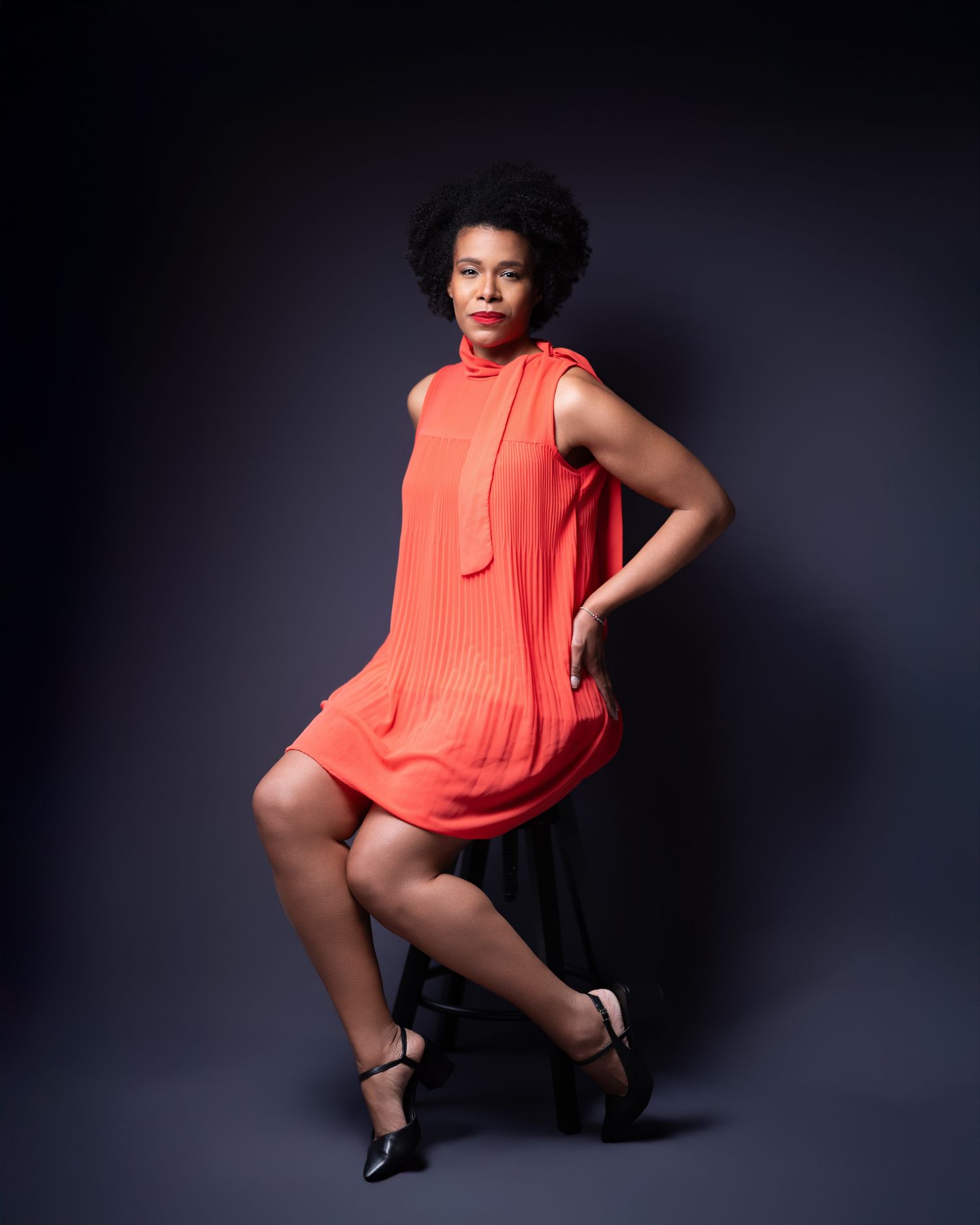 Colour studio portrait of a woman with natural hair in an orange halterneck dress, dark background, dynamic pose
