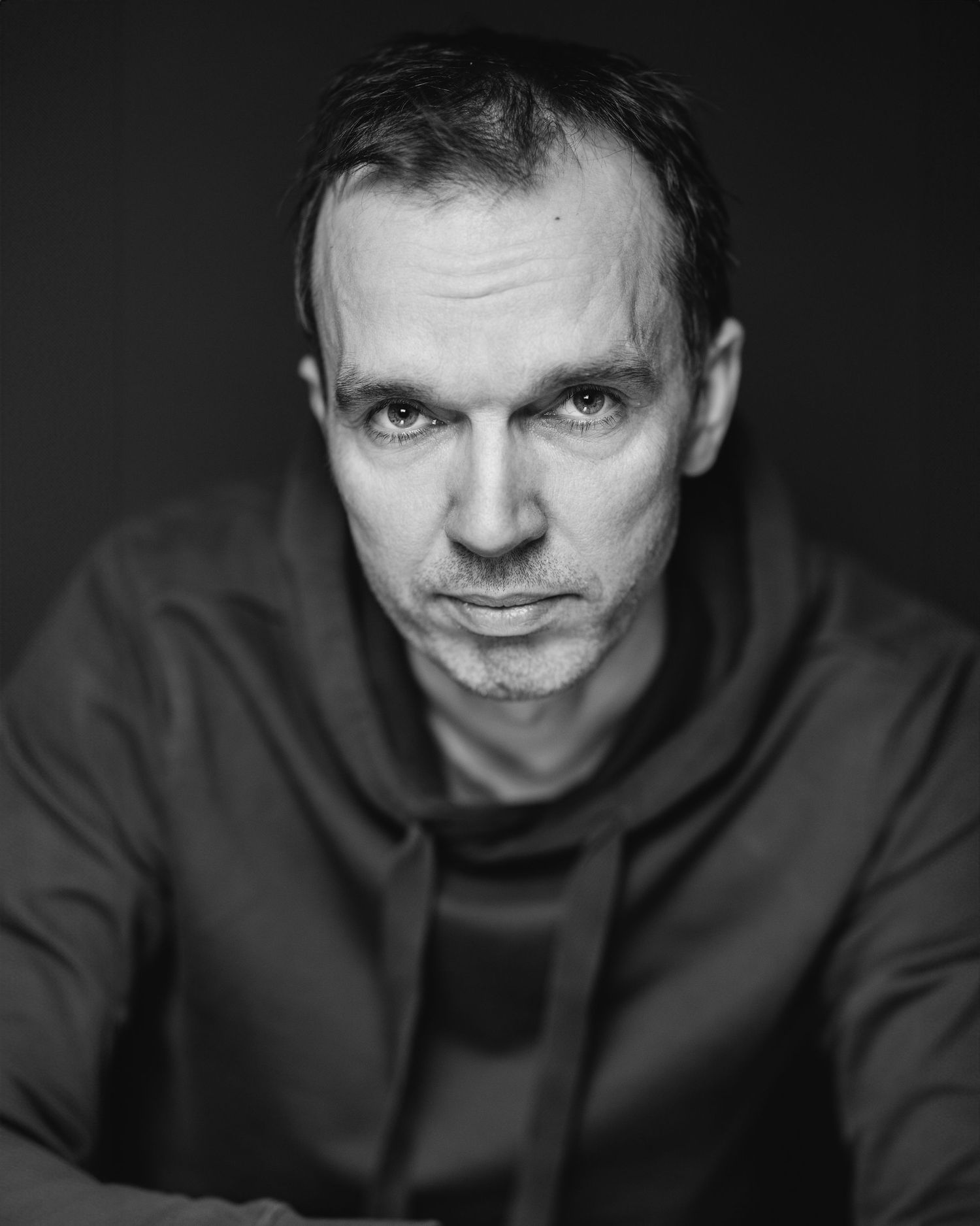 Black and white close-up portrait of a man with short hair in a dark hoodie, intense direct gaze, dark background