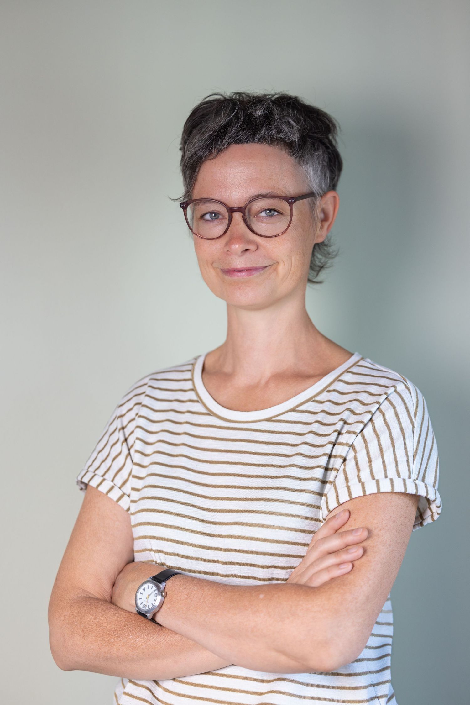 Colour portrait of a person with short dark hair and glasses in a striped t-shirt, arms crossed, light grey background