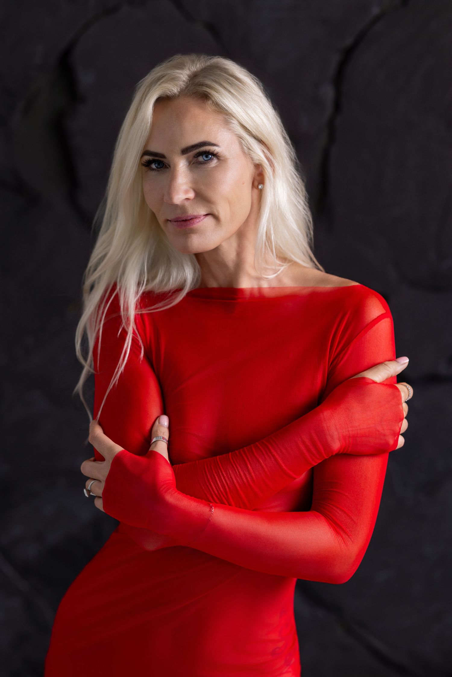 Studio portrait of a woman in a red long-sleeved dress, arms crossed, against a dark textured background