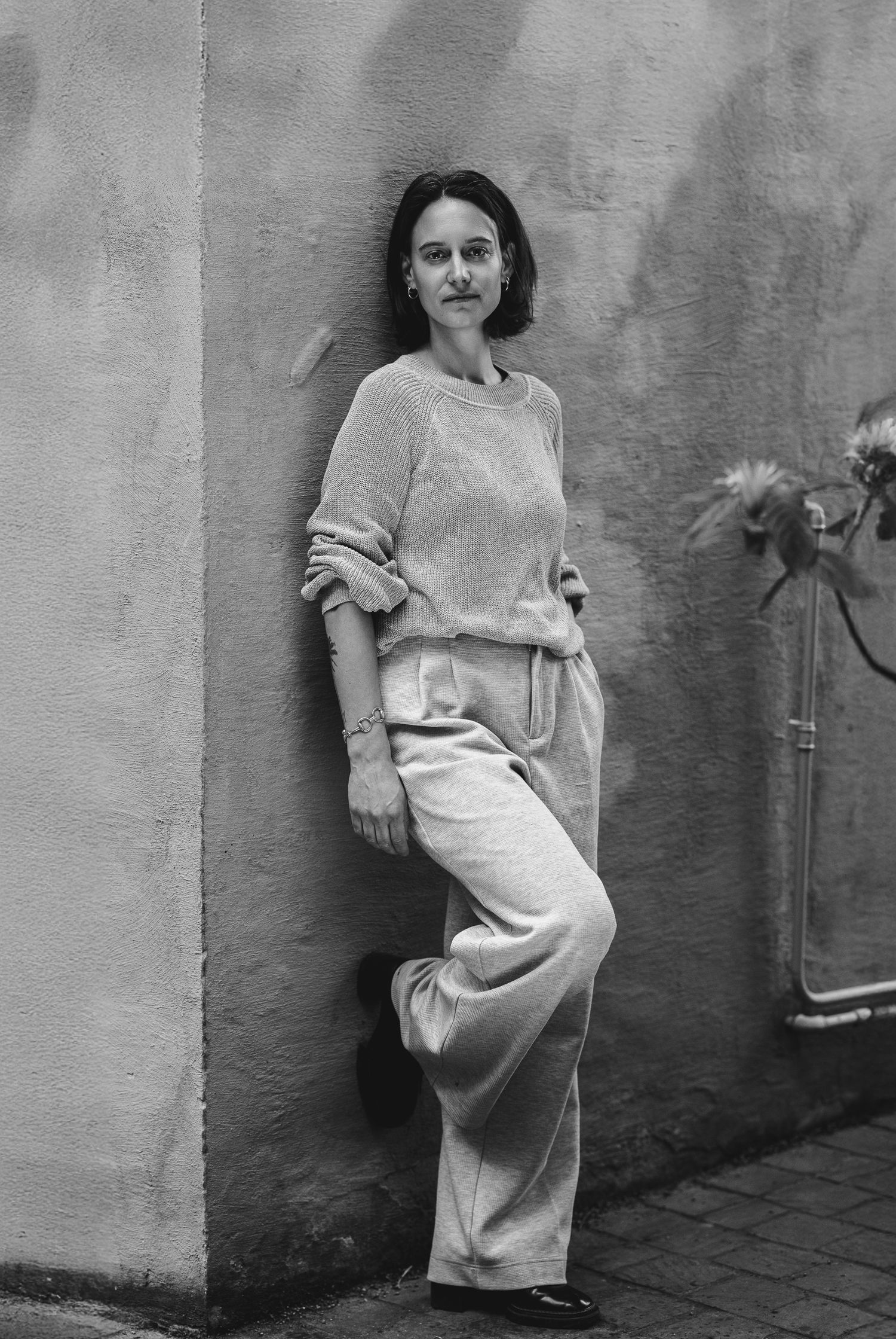 Black and white portrait of a woman in wide-leg trousers and a loose top, leaning against a plain wall, relaxed pose