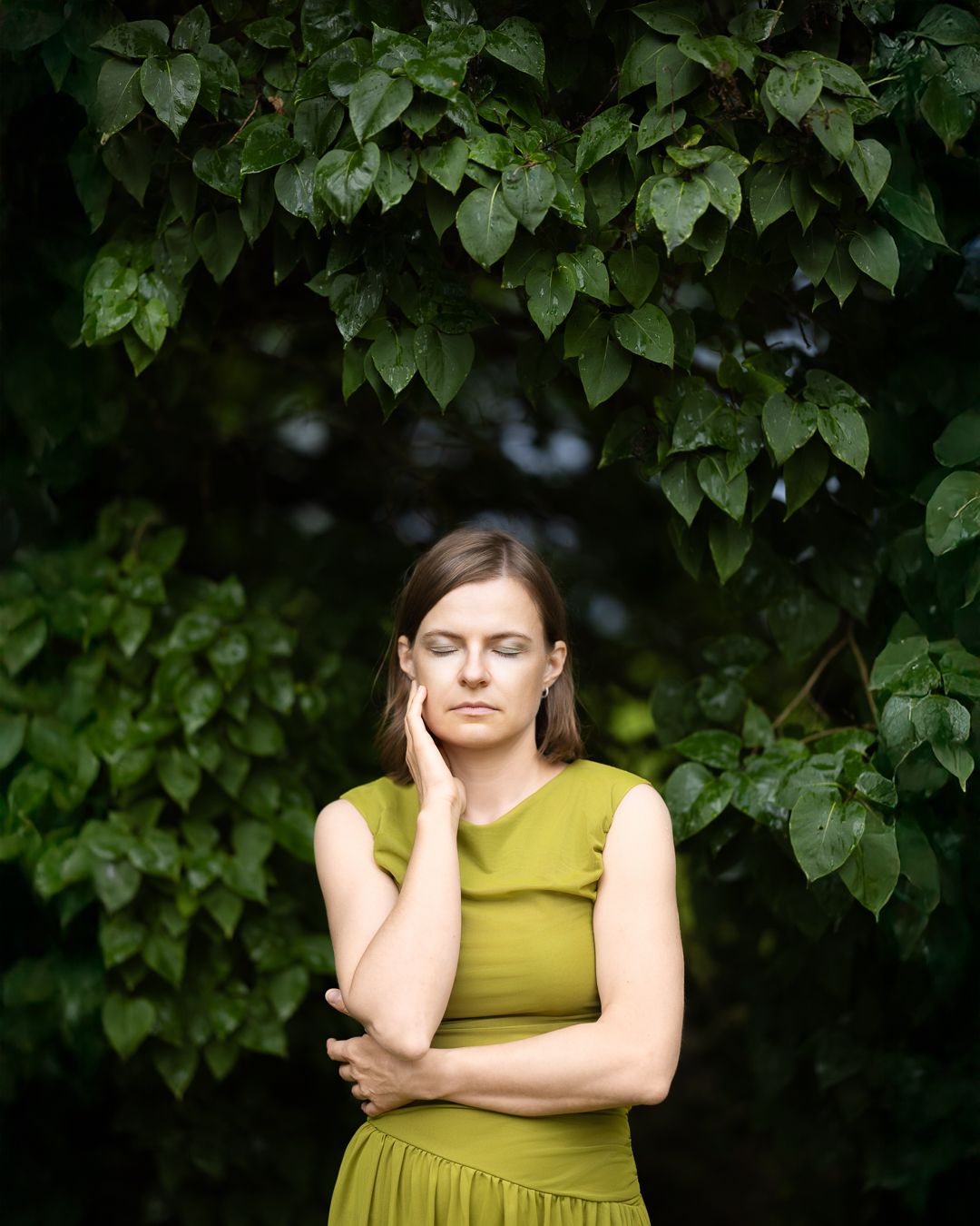 Colour portrait of a woman in an olive dress, eyes closed, hand to cheek, against dark ivy