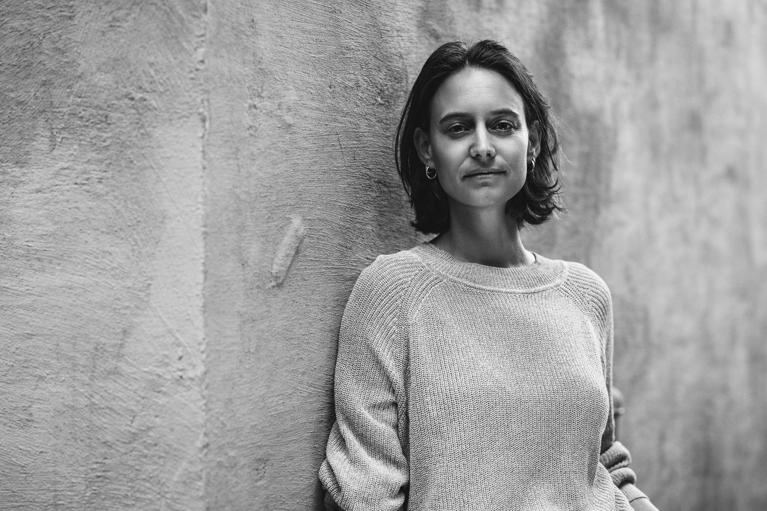 Black and white portrait of a woman in a light knit jumper, leaning against a textured concrete wall