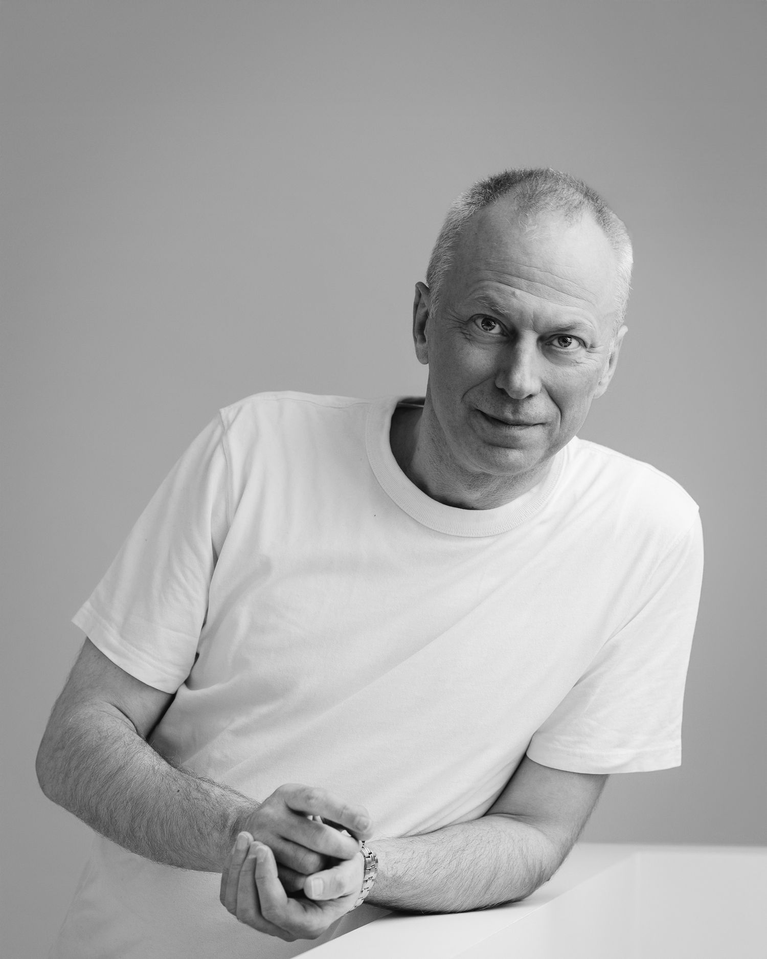 Black and white portrait of a man in a white t-shirt, leaning forward with hands clasped, light grey background