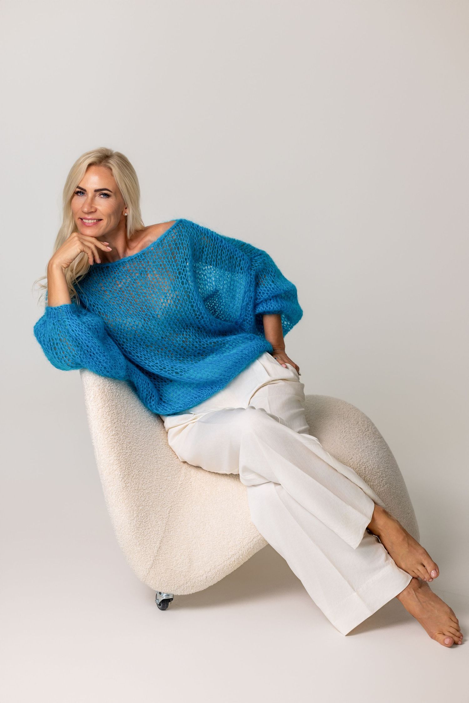 Colour portrait of a woman with long blonde hair in a sheer blue top and white trousers, seated on a chair, neutral background