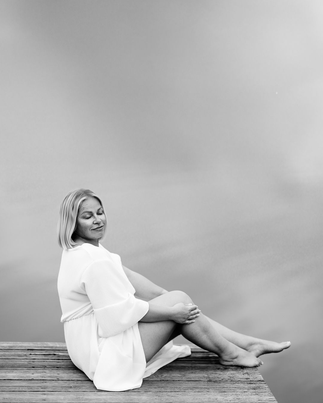 Black and white portrait of a woman in a white outfit seated on a wooden jetty, legs extended, serene composition