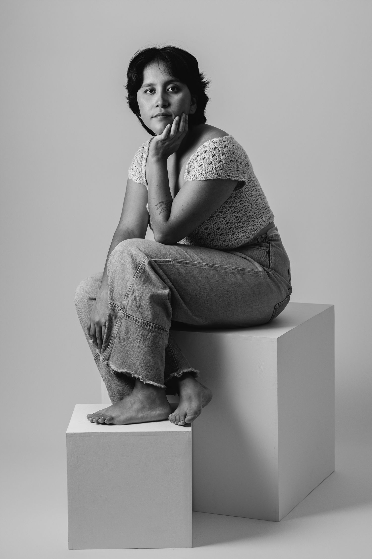 Black and white studio portrait of a woman in a lace top and wide-leg trousers, seated on a white cube, hand to chin
