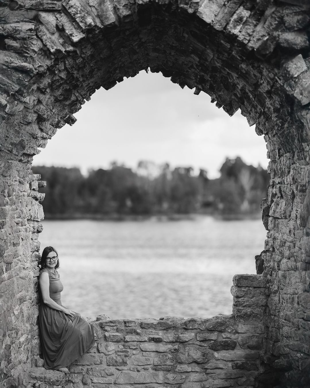 Black and white portrait of a woman seen through a large stone arch opening in ancient ruins, countryside behind