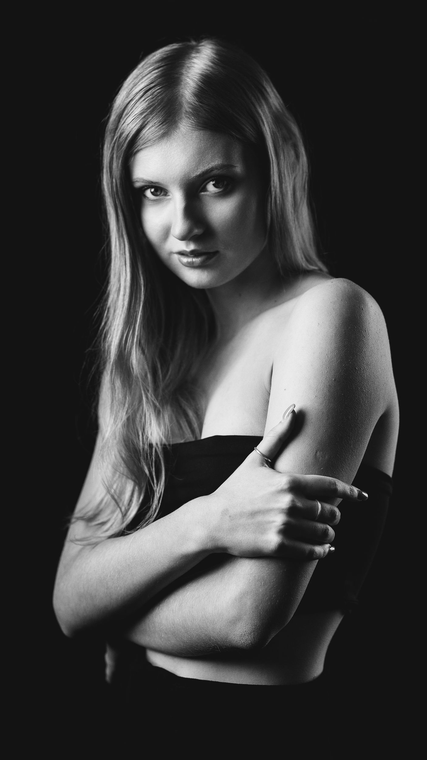 Black and white boudoir portrait of a woman with long blonde hair, arms crossed, against a dark background, direct gaze
