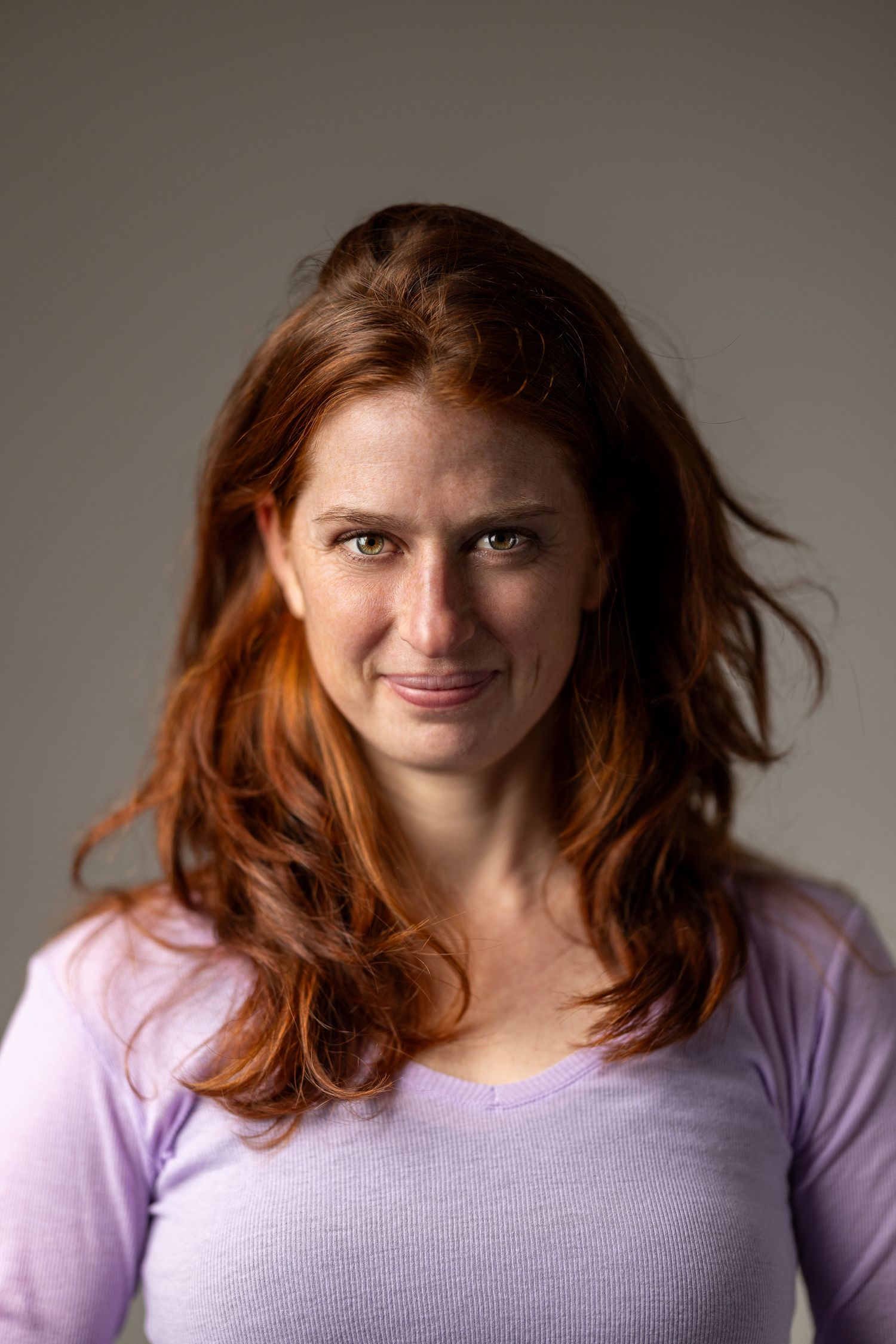 Colour portrait of a woman with auburn hair in a lavender long-sleeve top against a grey background