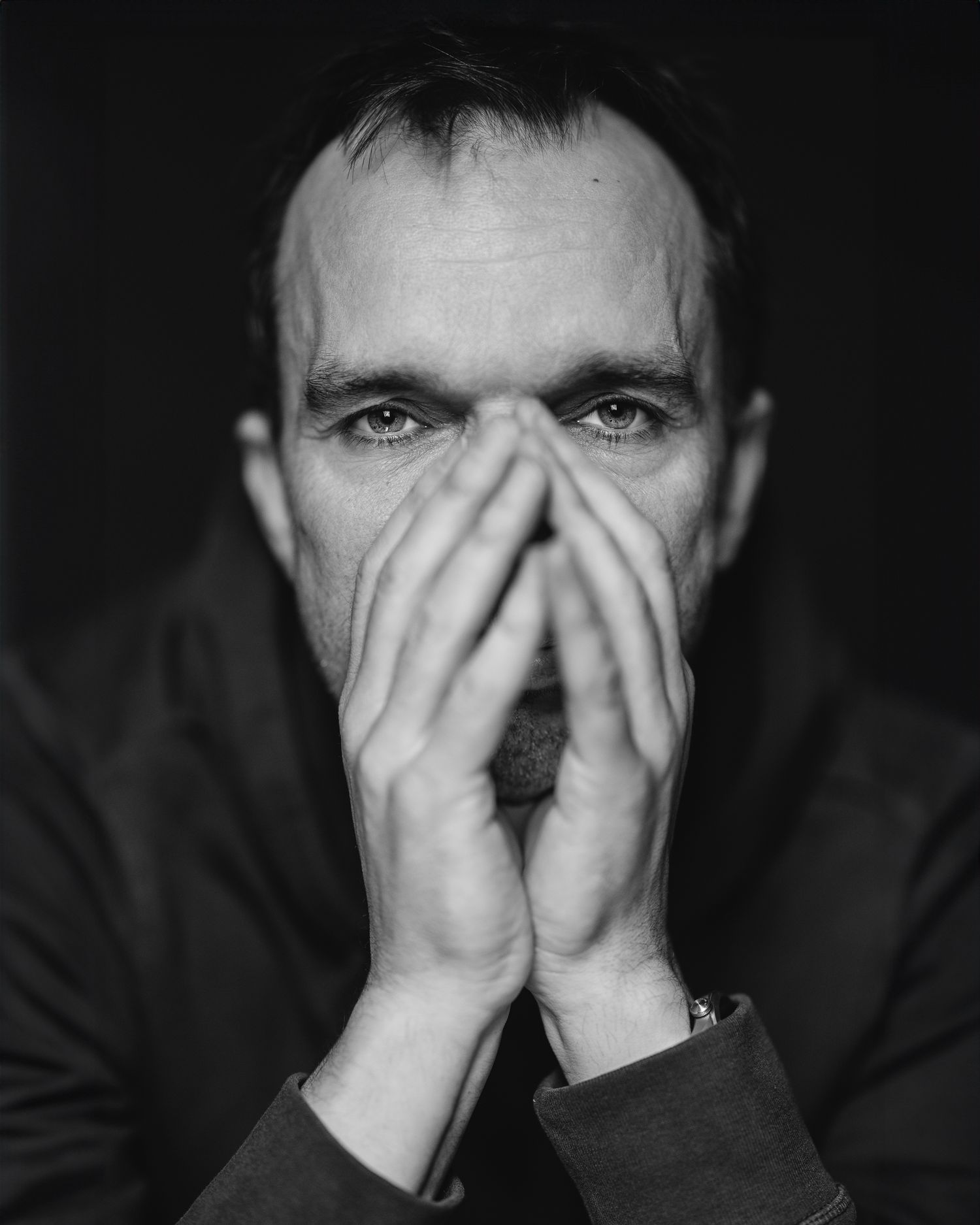 Black and white close-up portrait of a man in a dark suit, hands pressed together over his mouth, intense gaze