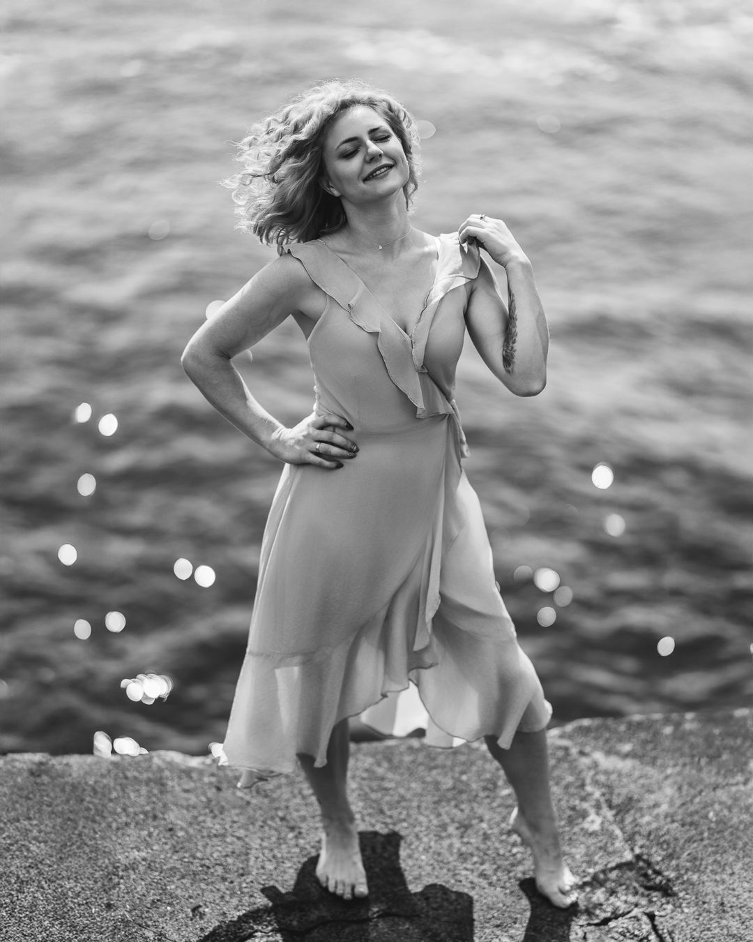Black and white portrait of a woman in a long skirt standing on coastal rocks beside the sea, arms extended